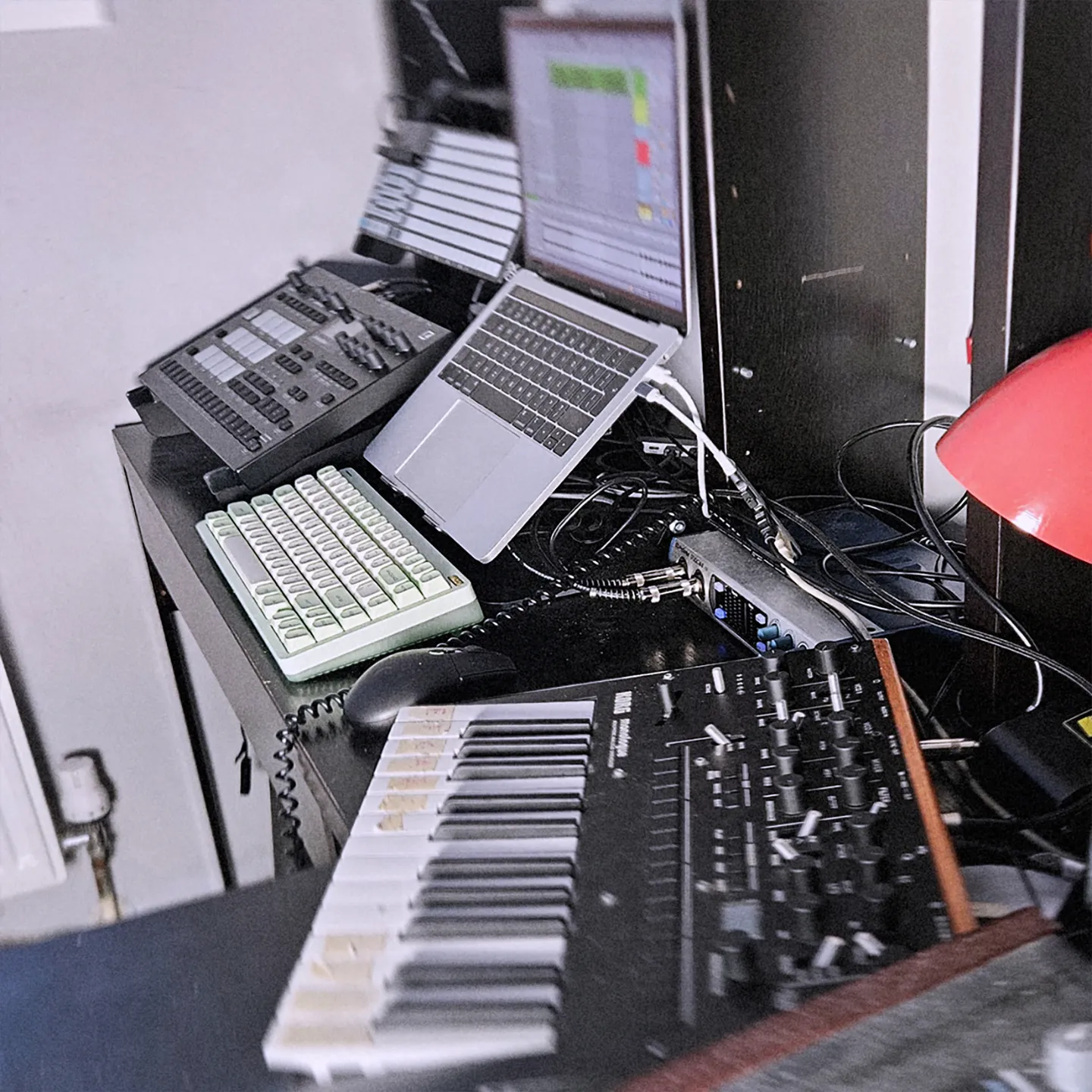 CO:LOG's studio desk with a laptop running a DAW, a Korg synth, a green mechanical keyboard, an audio interface and a red lamp.