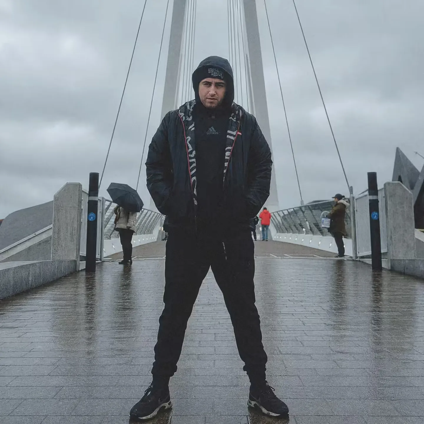 Jay-Lee stands on a pedestrian bridge in a black hooded jacket and beanie, hands in his pockets, on an overcast day.
