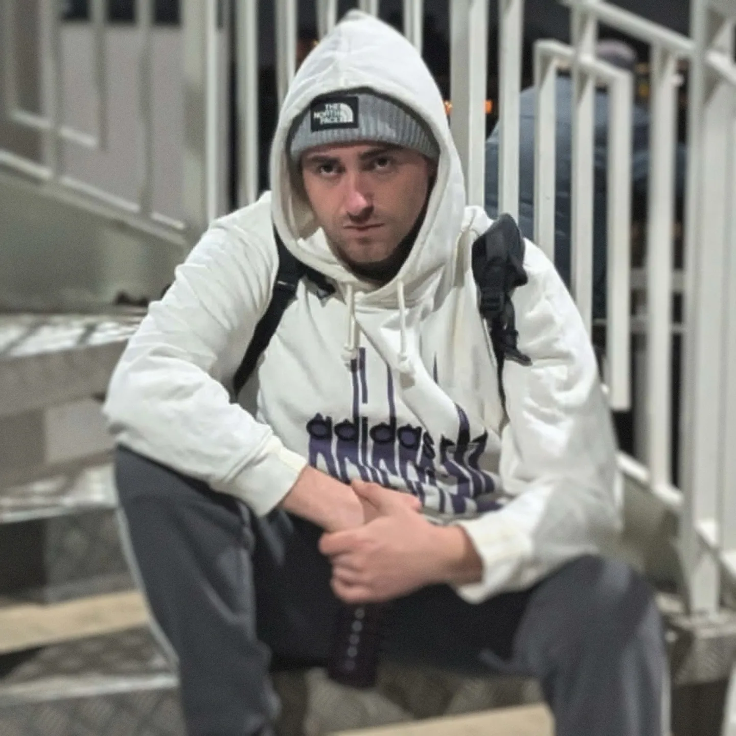 Jay-Lee sits on an outdoor staircase in a white hoodie and beanie, resting his arms on his knees.
