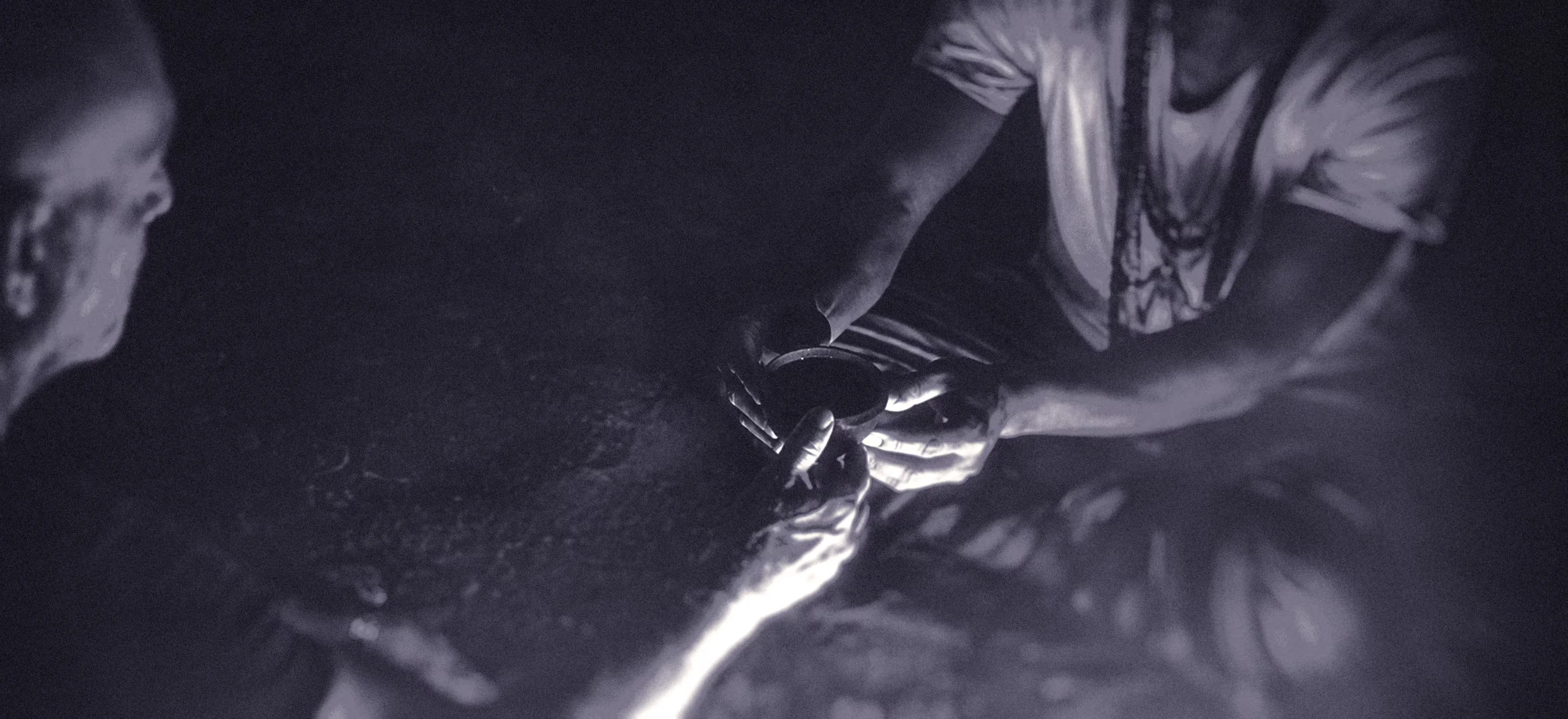 Two people are sitting in low light at an ayahuasca ritual, with one person handing a small round bowl, to the other. The image is for an article about self-doubt as a music producer.