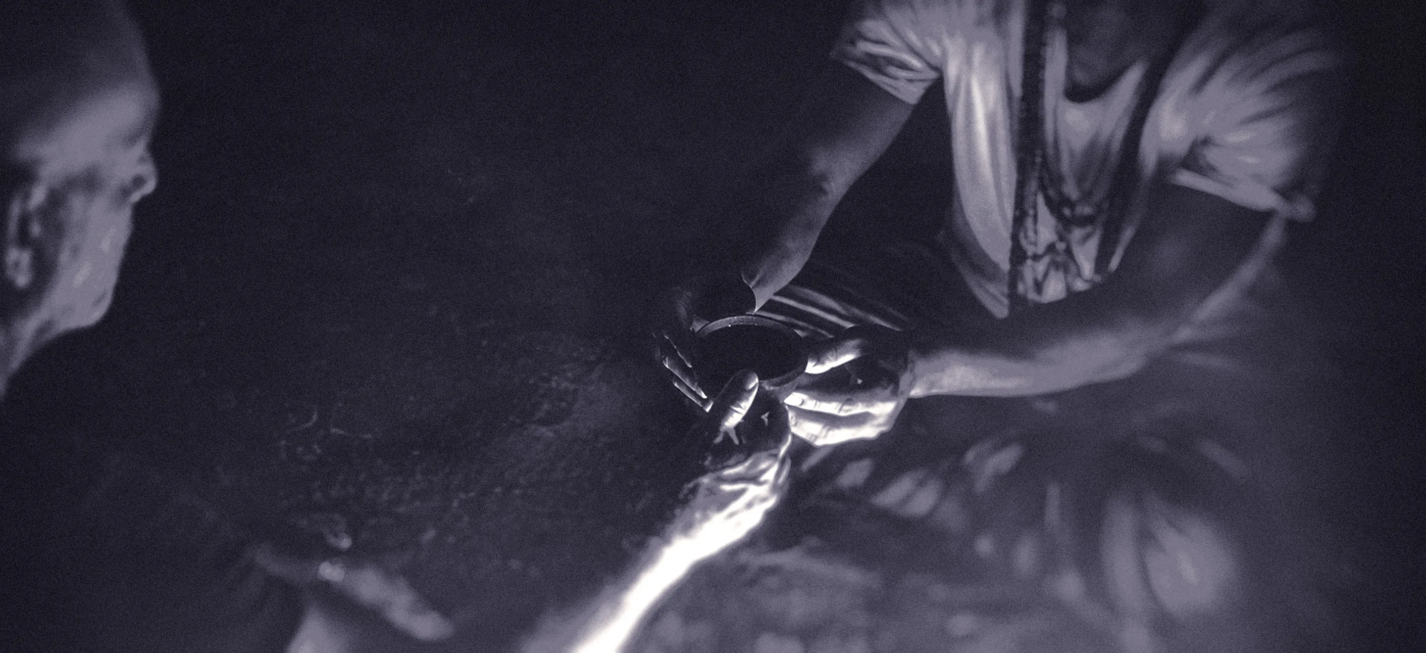 Two people are sitting in low light at an ayahuasca ritual, with one person handing a small round bowl, to the other. The image is for an article about self-doubt as a music producer.