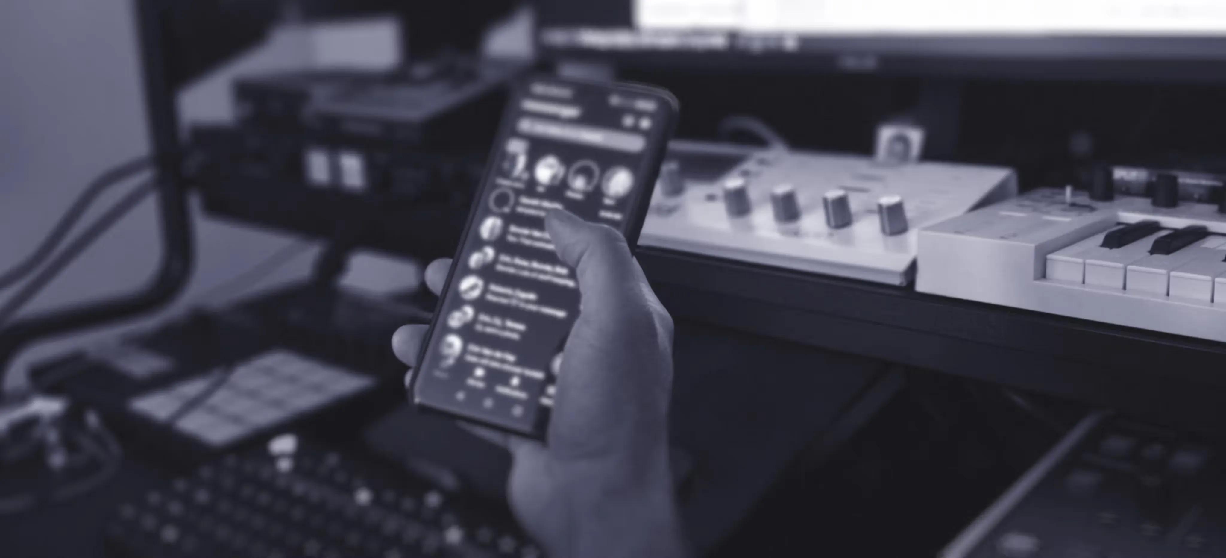 A person holds a smartphone displaying a messaging app in front of a music production setup with a keyboard, audio interface, and computer monitor. The scene gives a sense of distraction and lack of focus.
