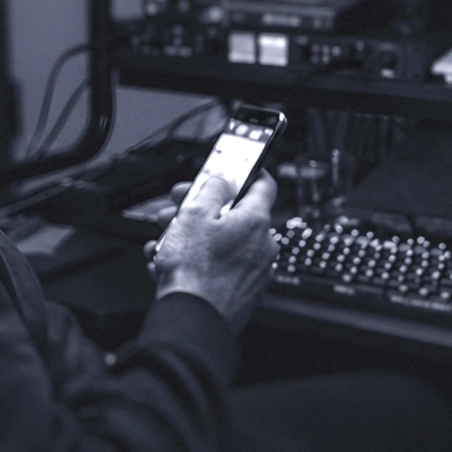 A man is seated at a computer, with studio equipment in the background, engaged with his cell phone in hand.
