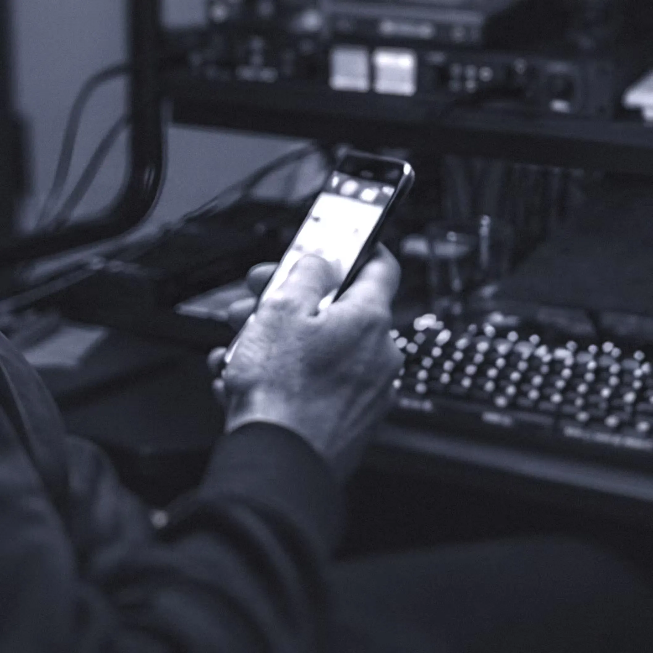 A man is seated at a computer, with studio equipment in the background, engaged with his cell phone in hand.
