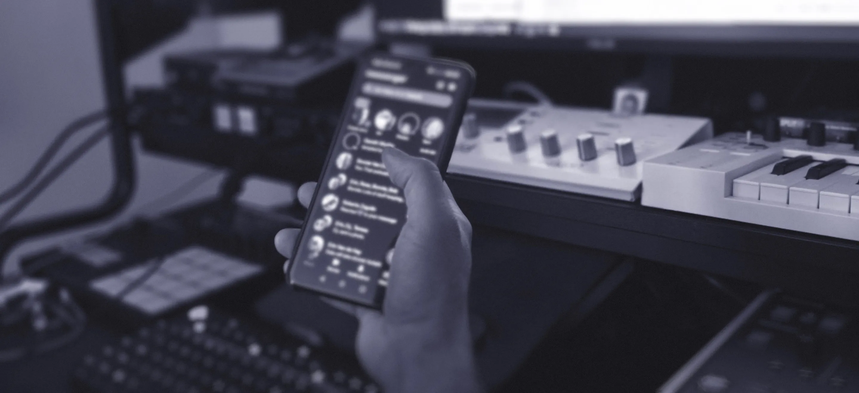 A person holds a smartphone displaying a messaging app in front of a music production setup with a keyboard, audio interface, and computer monitor. The scene gives a sense of distraction and lack of focus.