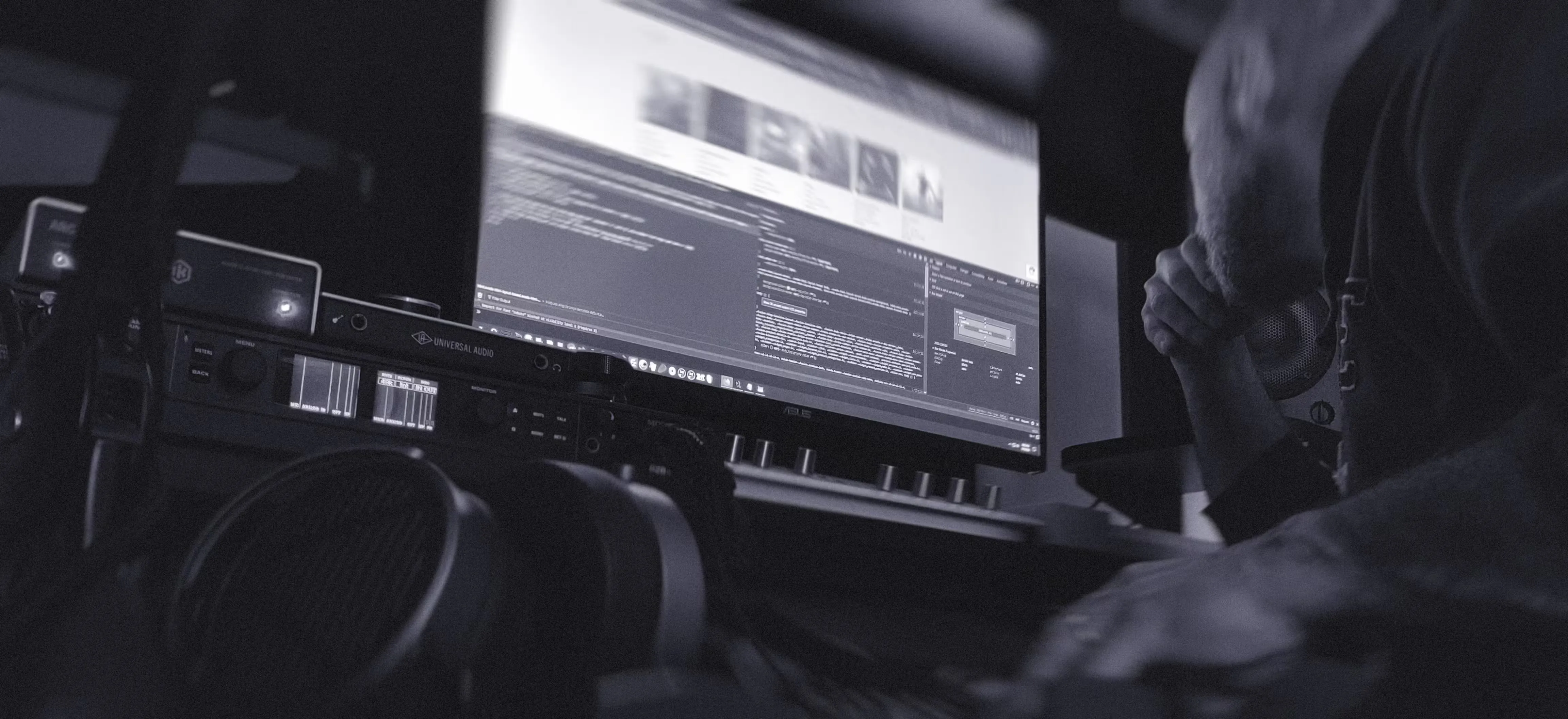 A person leans over a desk, surrounded by audio equipment, looking at a large computer monitor displaying code and images in a dimly lit workspace, capturing a moment of creative development for music artists.