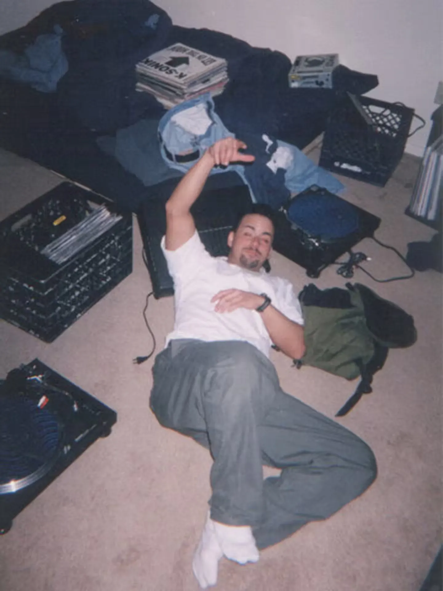 A man lies on the carpeted floor, surrounded by vinyl records, a turntable, milk crates, and an unmade bed in a messy bedroom.