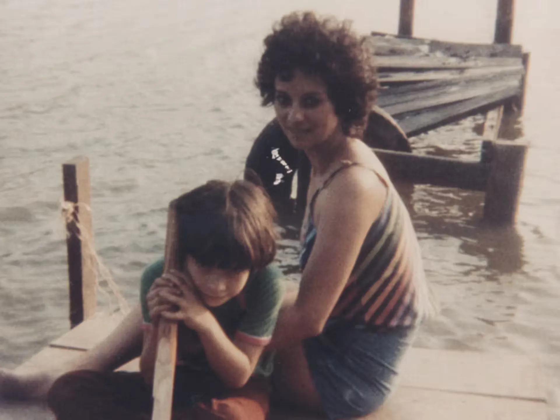 A woman and a child sit on a wooden dock by the water. The woman is wearing a striped top and shorts, while the child leans on a wooden post. A damaged dock is visible in the background.