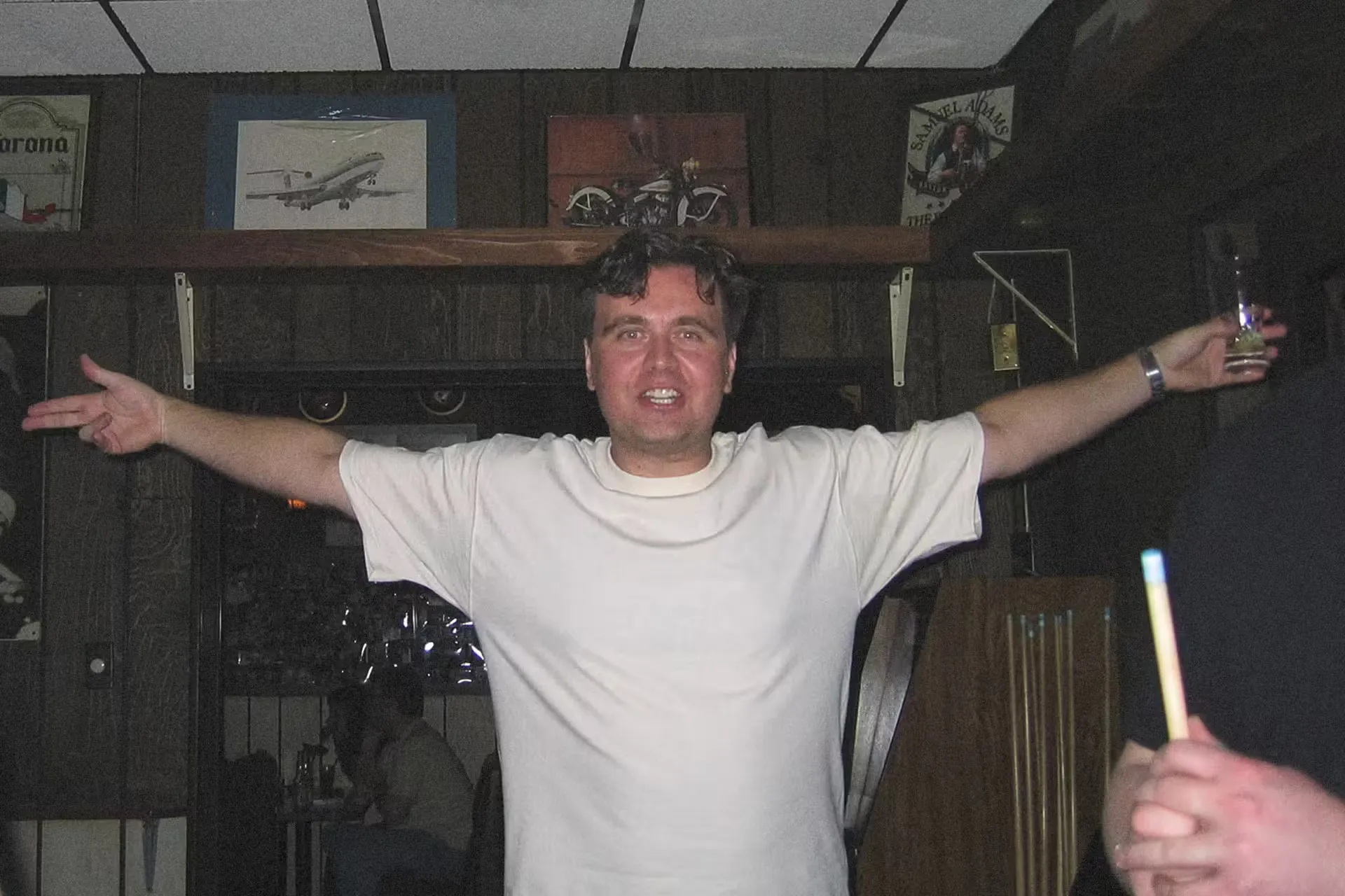 A smiling man stands in a dimly lit bar with his arms open wide. He wears a white shirt, exuding a welcoming and cheerful vibe.