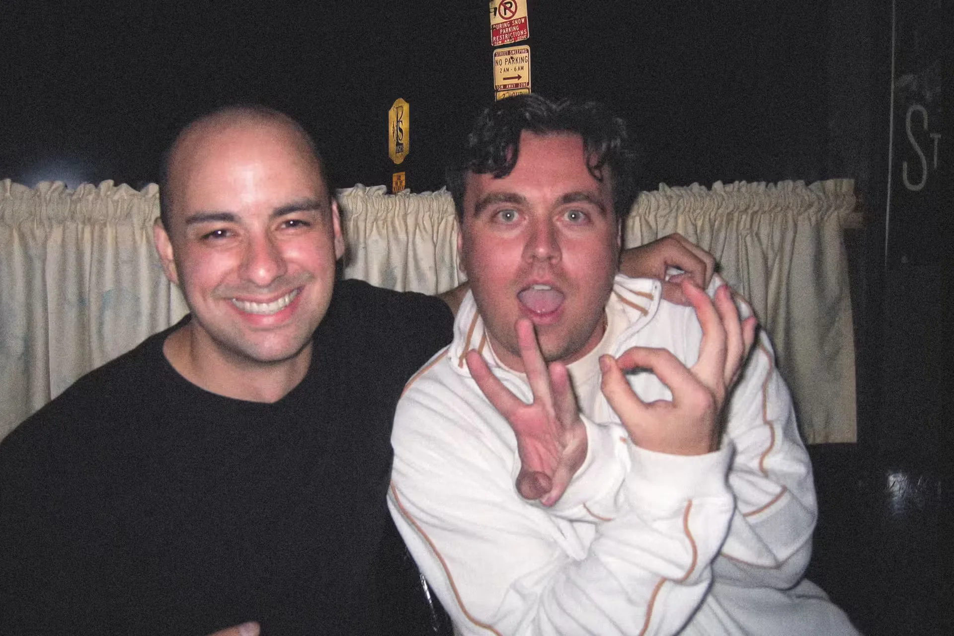 Two men pose playfully in a dimly lit room with curtains in the background. The man on the left smiles broadly, while the man on the right makes a hand gesture.