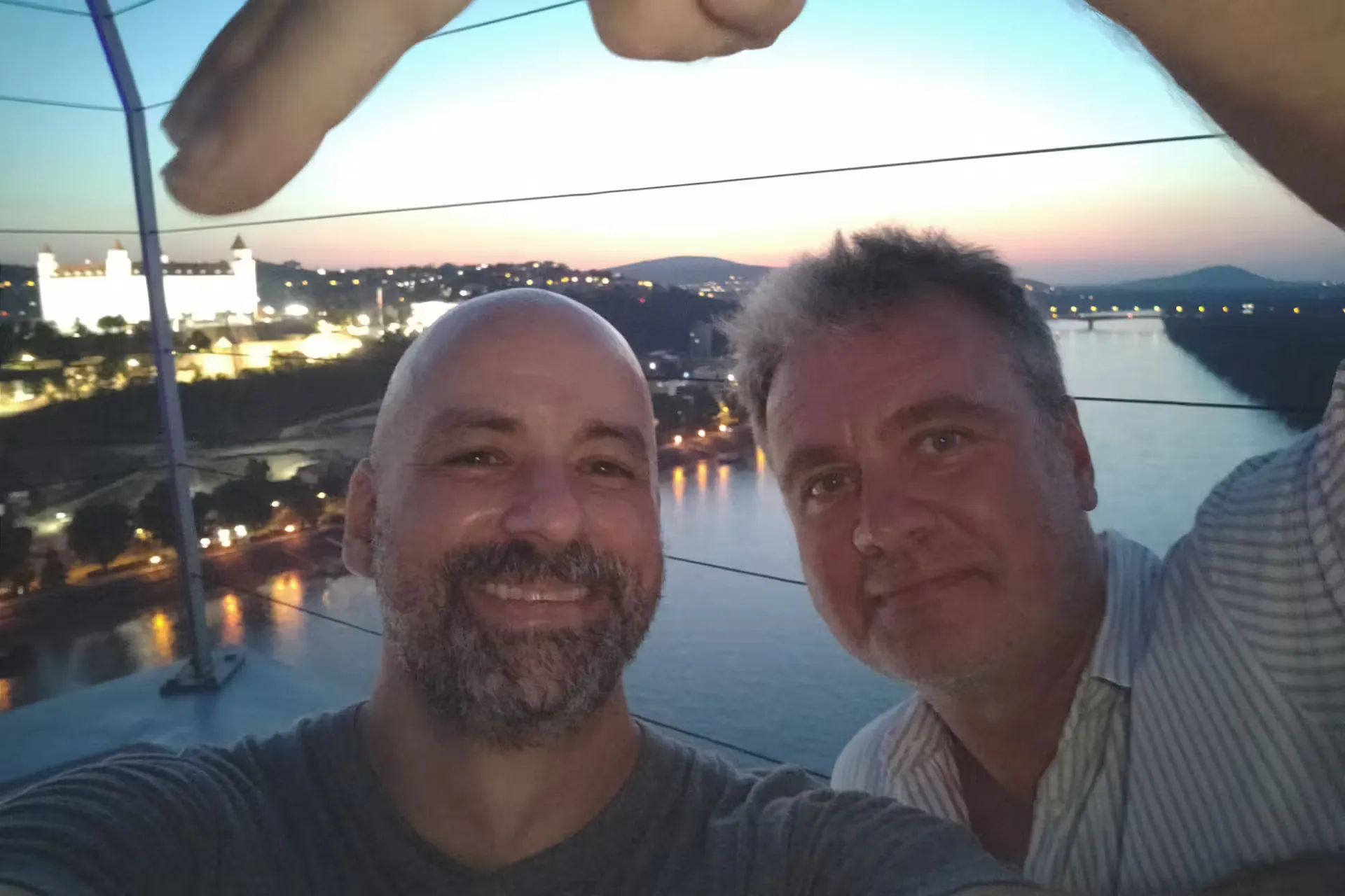 Two men smiling in front of a river at dusk, with a cityscape and a lit castle in the background. The atmosphere is relaxed and cheerful.