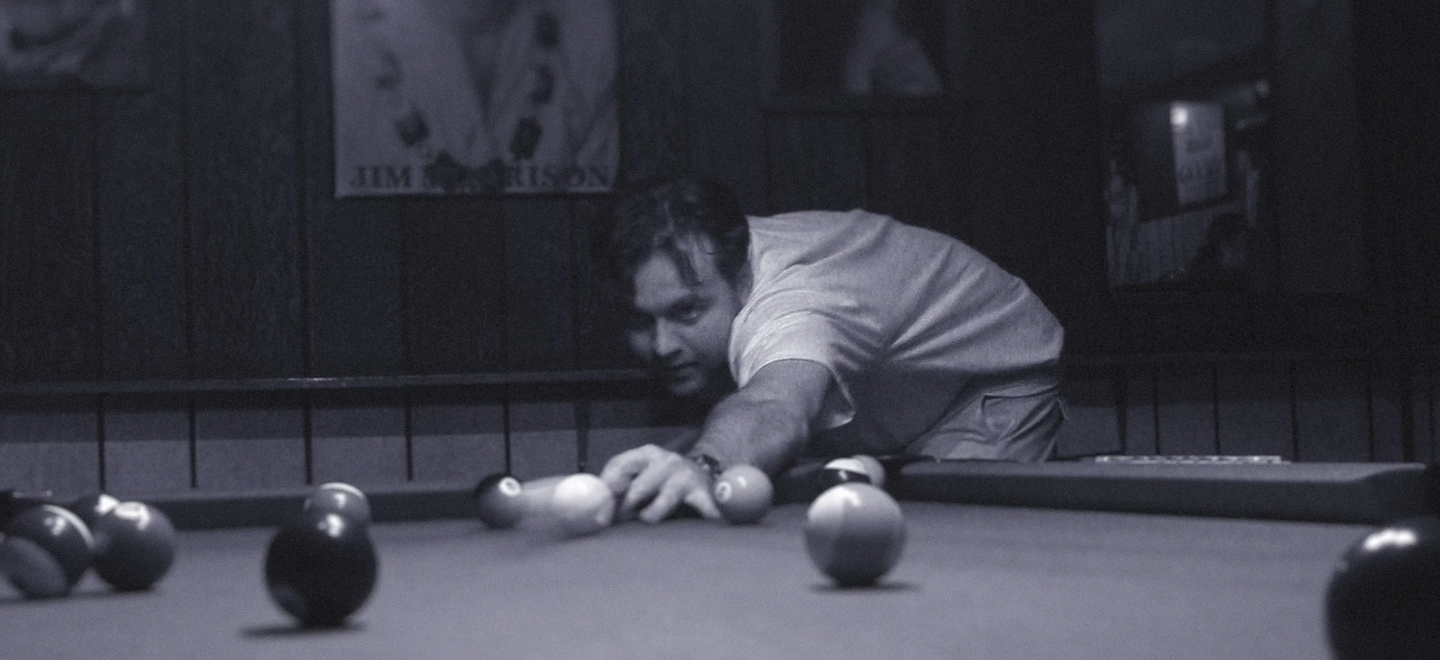 A man intensely focuses on a pool shot in dim lighting, surrounded by billiard balls. The serene atmosphere suggests concentration and leisure.