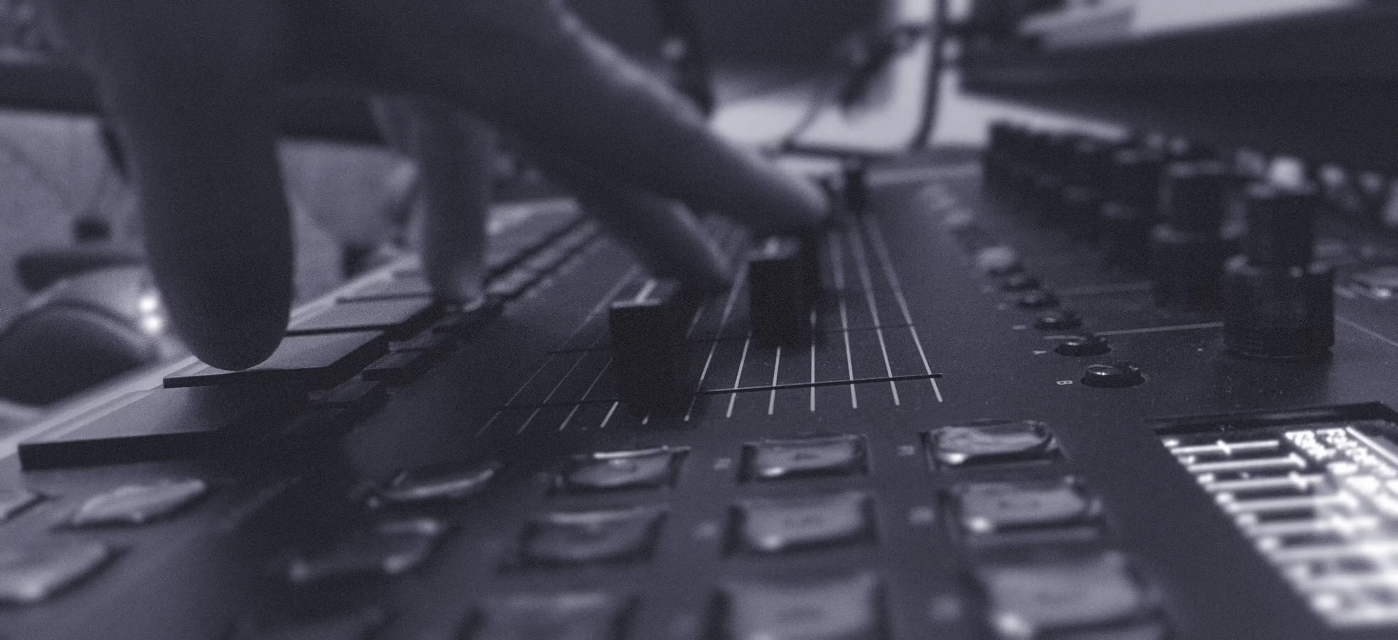 A producer coaching with hand adjusting sliders on a sound mixing console, control knobs and buttons are visible in the foreground.