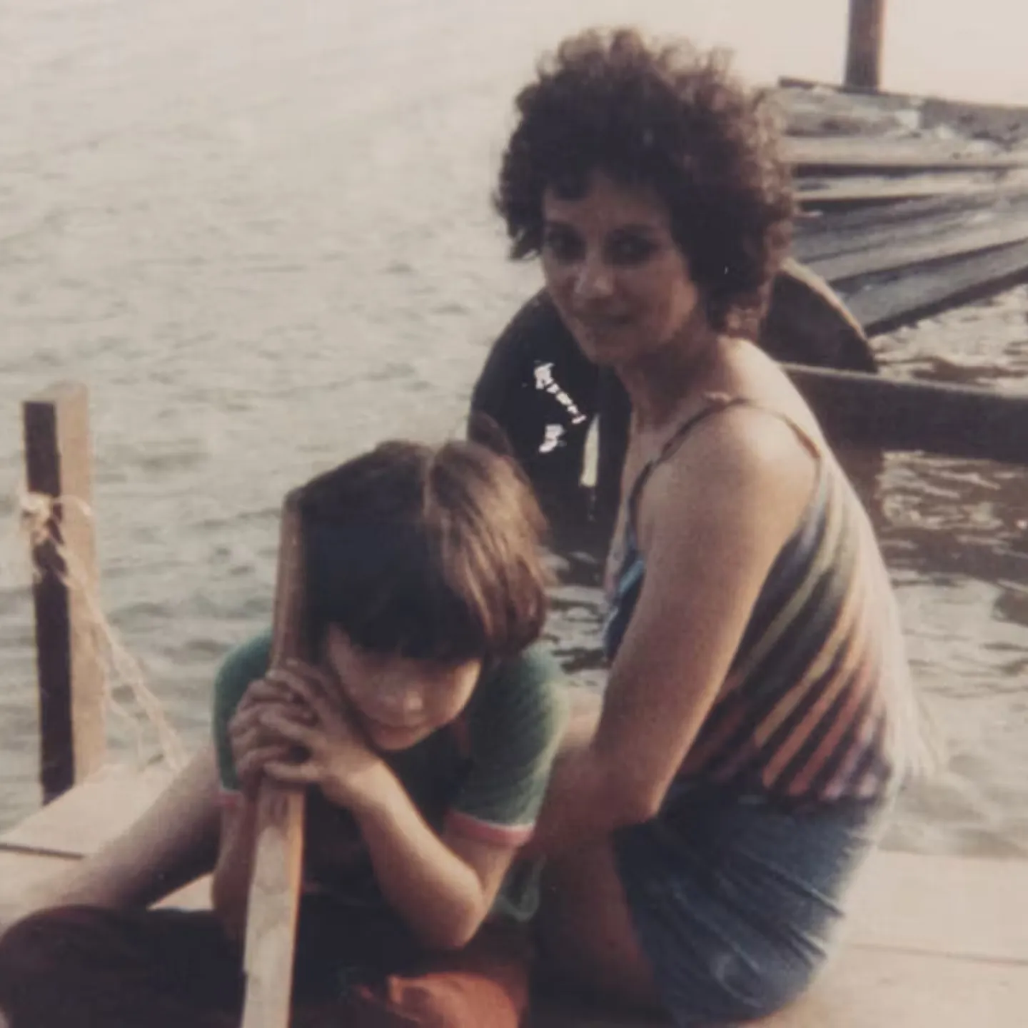 A woman and a child sit on a wooden dock by the water. The woman is wearing a striped top and shorts, while the child leans on a wooden post. A damaged dock is visible in the background.