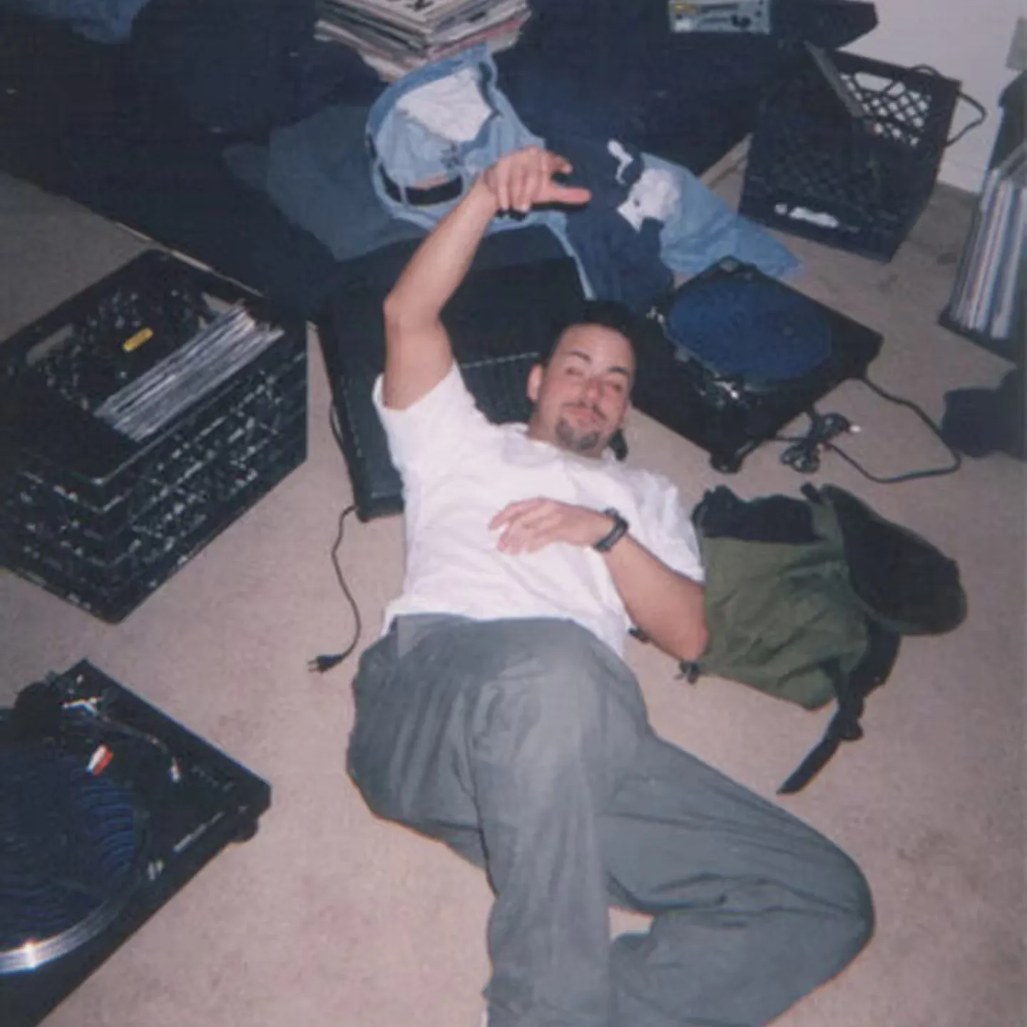 A man lies on the carpeted floor, surrounded by vinyl records, a turntable, milk crates, and an unmade bed in a messy bedroom.