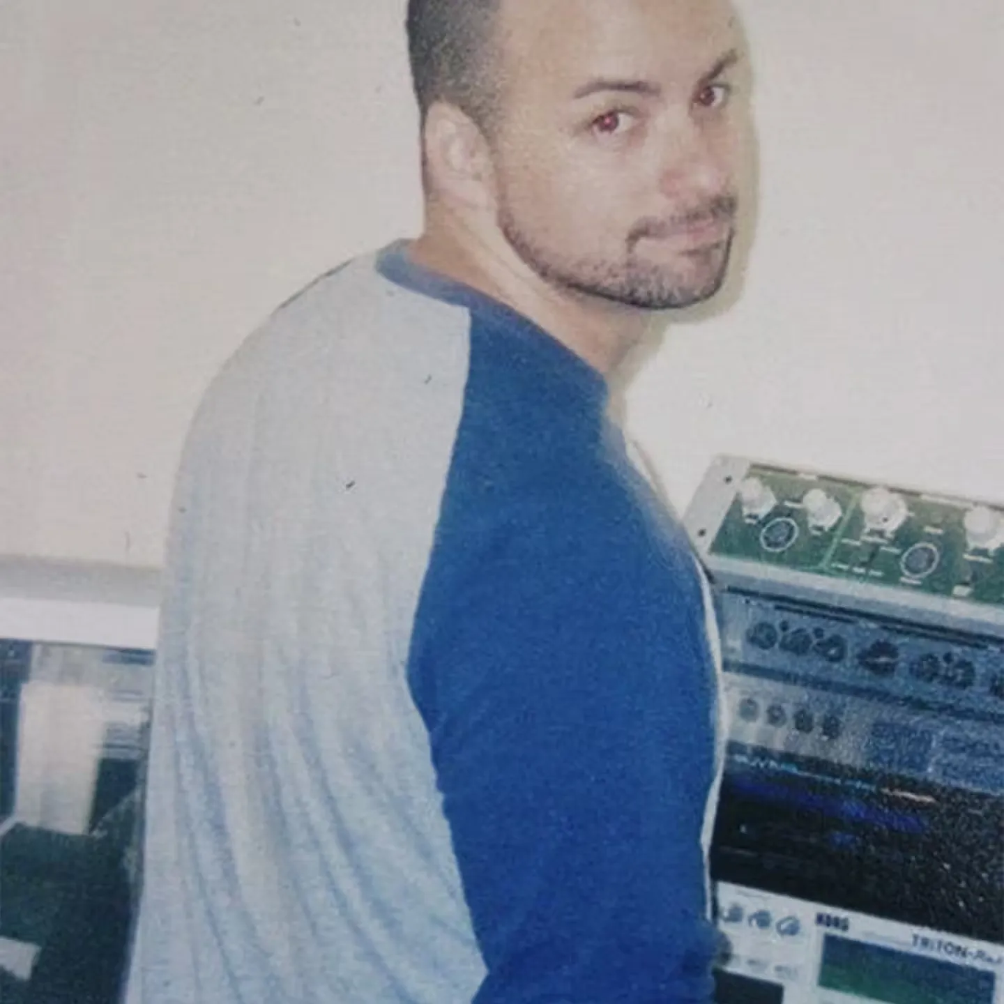 A man with a shaved head and trimmed beard, wearing a gray and blue shirt, stands in front of electronic audio equipment, looking back over his shoulder.
