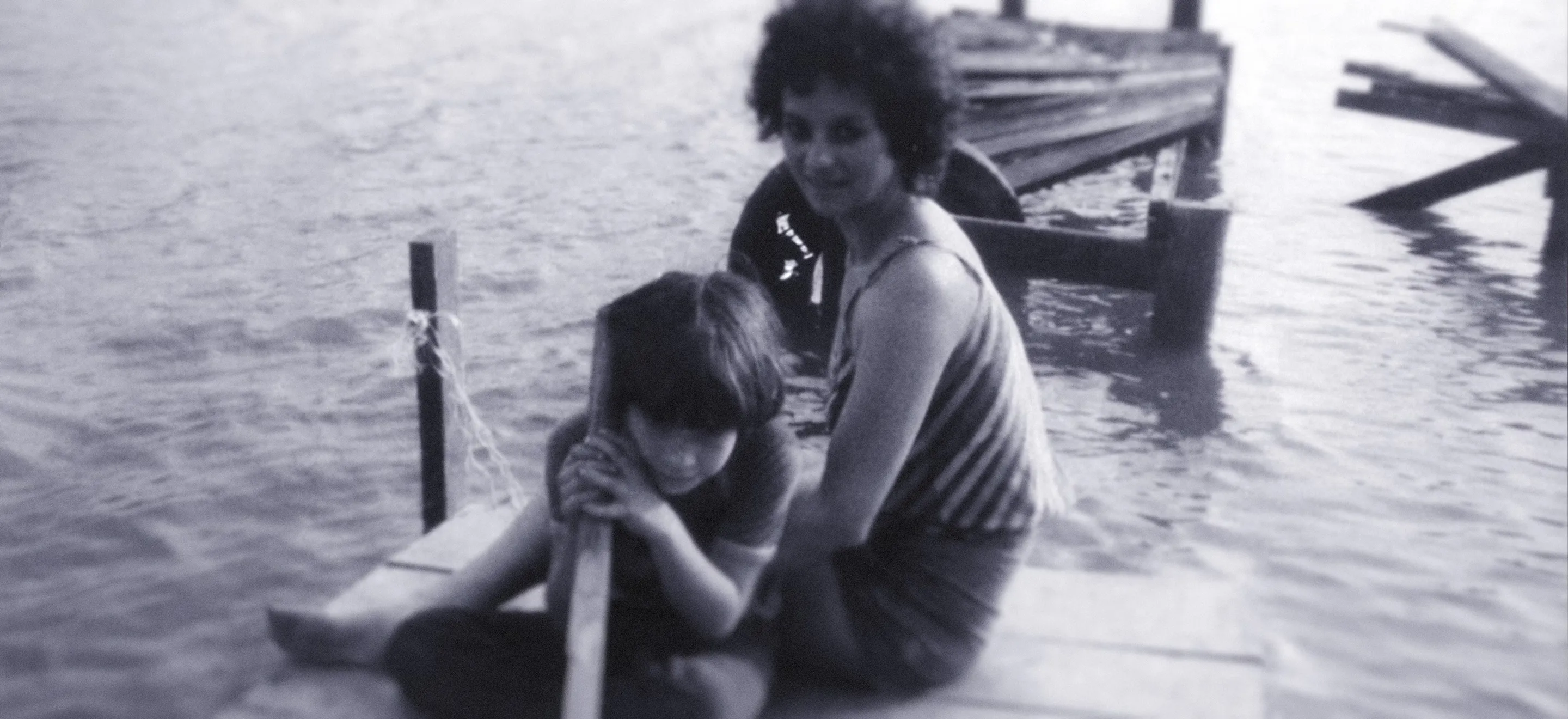 Monochrome photograph of a woman and a child sit on a wooden dock by the water; the child is holding a pole while the woman sits behind, both facing the camera.