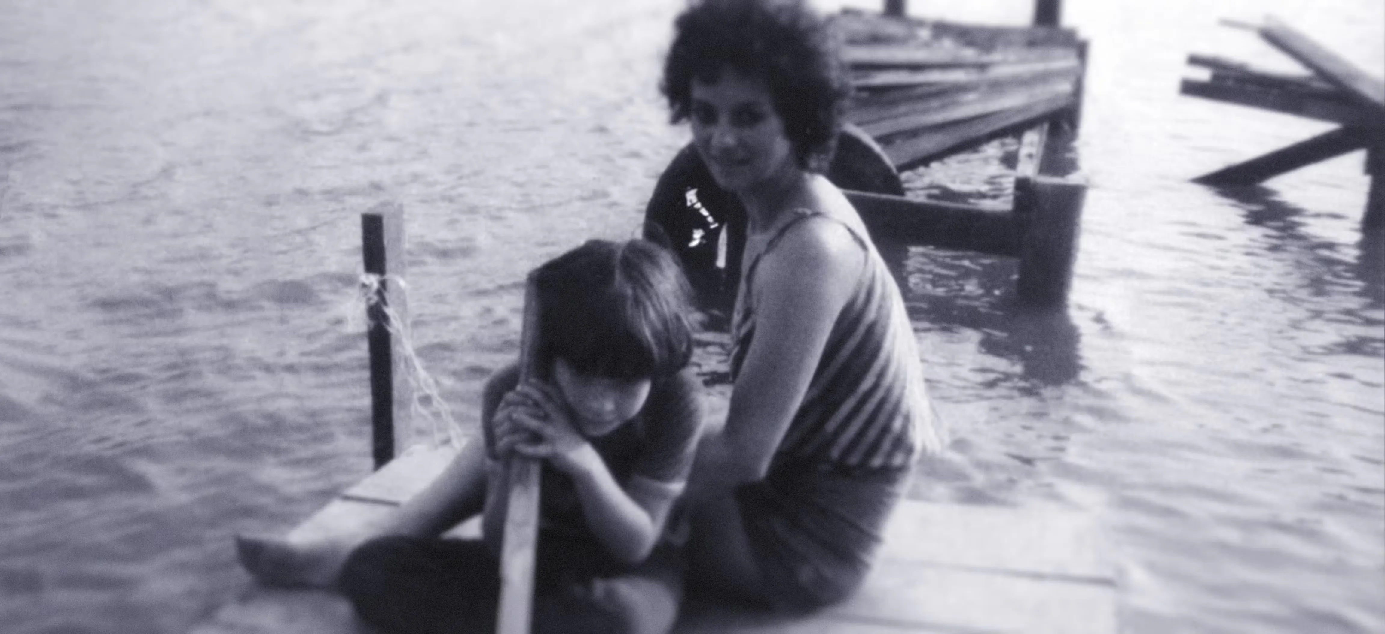 Monochrome photograph of a woman and a child sit on a wooden dock by the water; the child is holding a pole while the woman sits behind, both facing the camera.