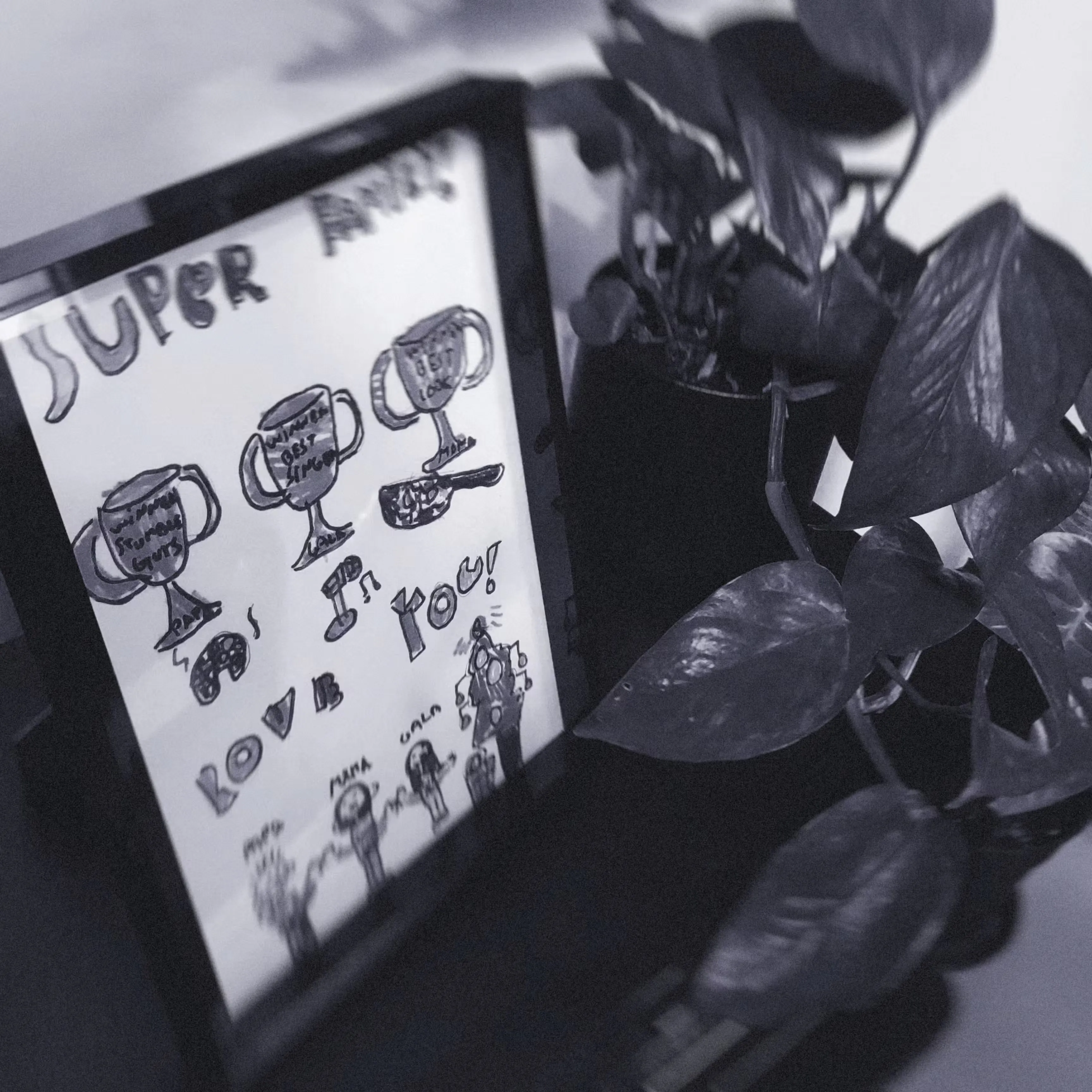 Framed child’s drawing of trophies with text "Super Family" and "I Love You!" next to a leafy plant, conveying encouragement and love.