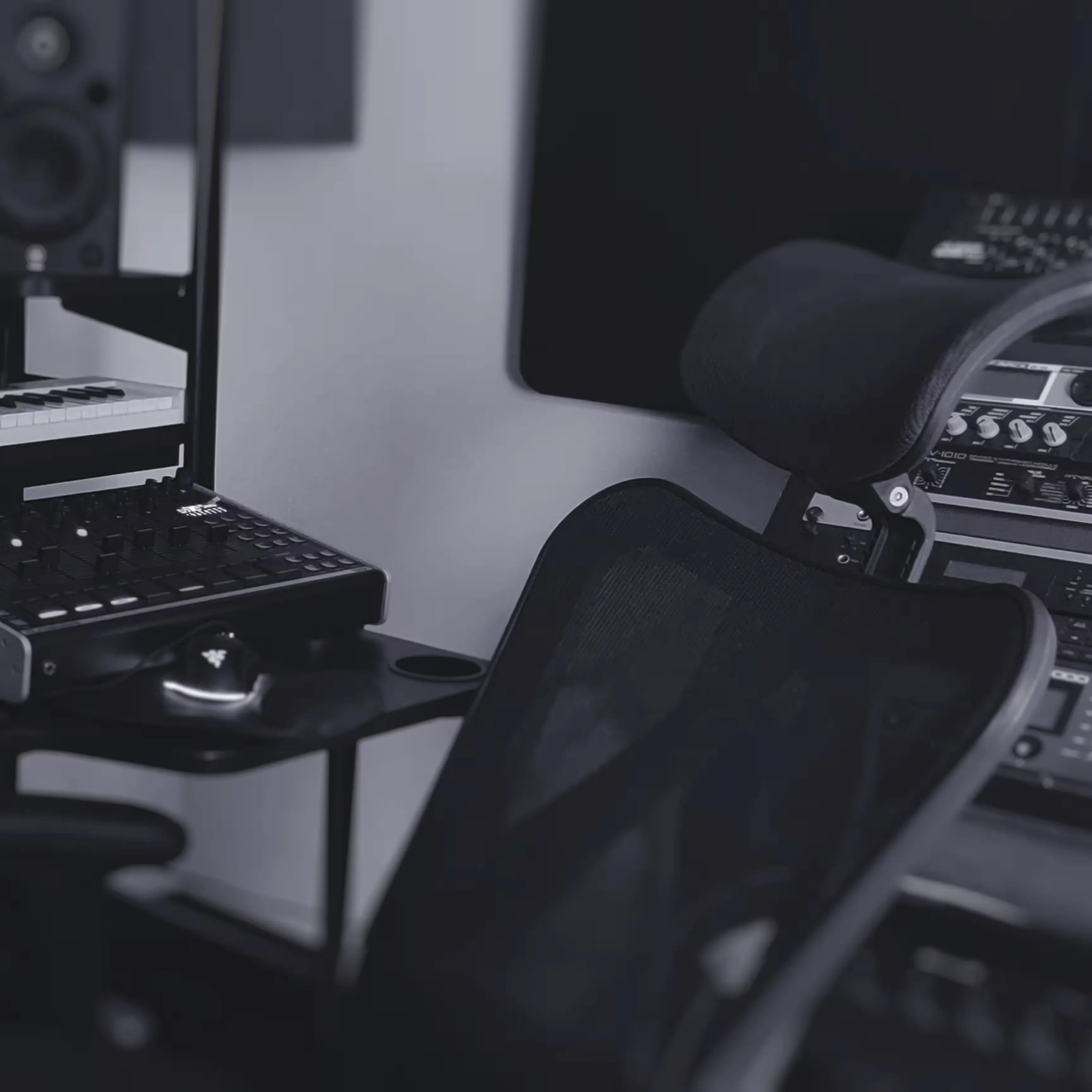 A black mesh office chair is positioned in front of audio equipment, including a mixer, speakers, and rack-mounted gear in a music studio.