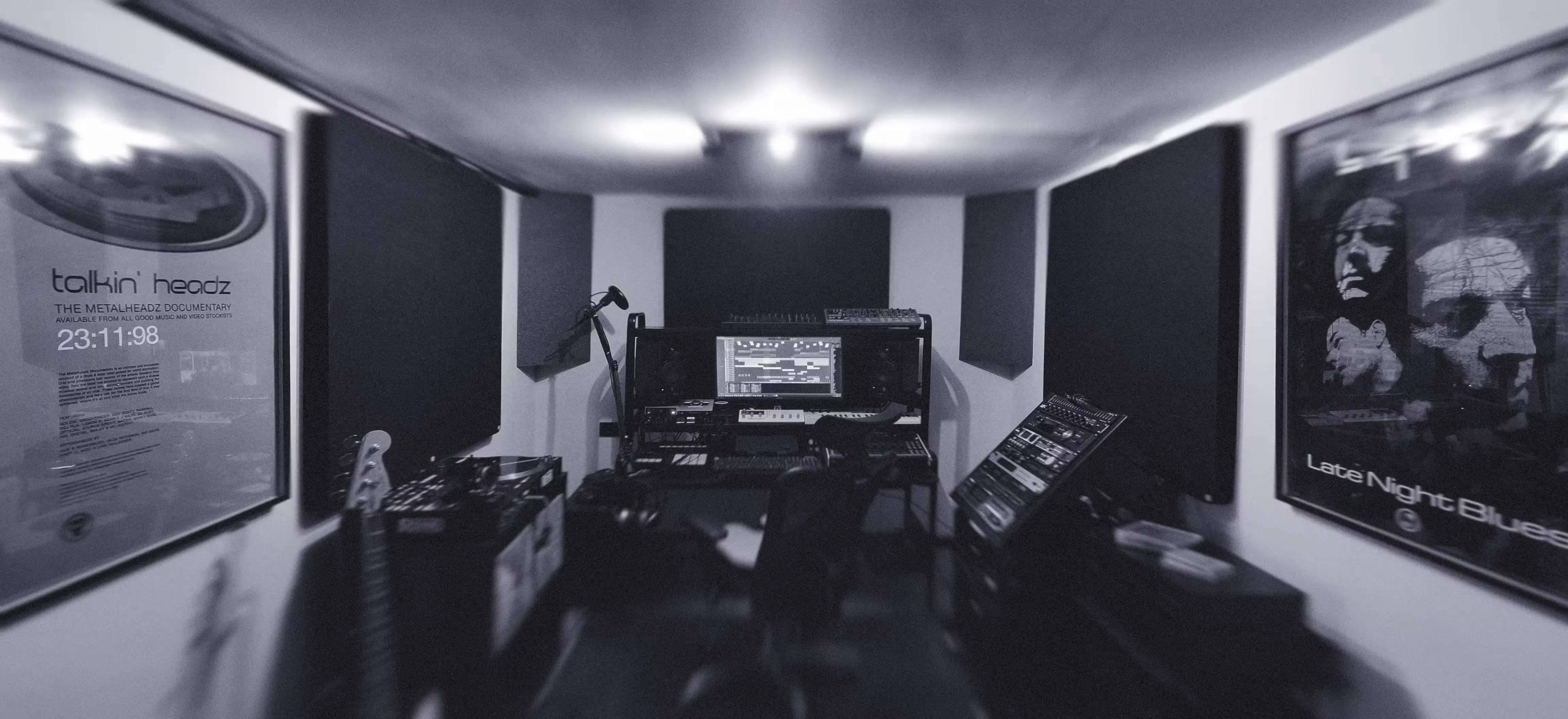 Monochrome photograph of a dimly lit music studio with sound panels, a computer on a desk, and various musical equipment. Posters adorn the walls, creating a focused yet creative vibe.