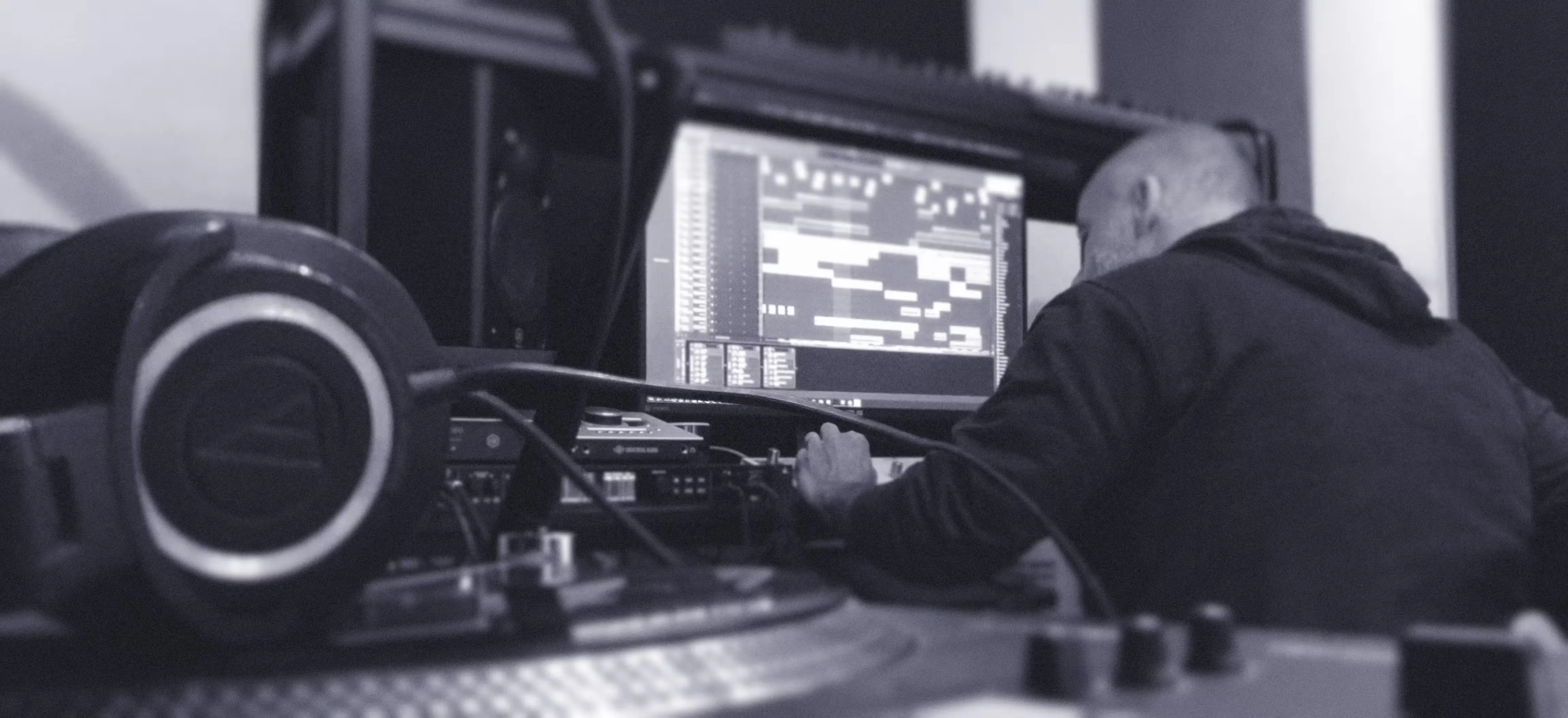 Monochrome photograph of a man working at a music production studio desk with headphones, mixer, and DAW software on screen.
