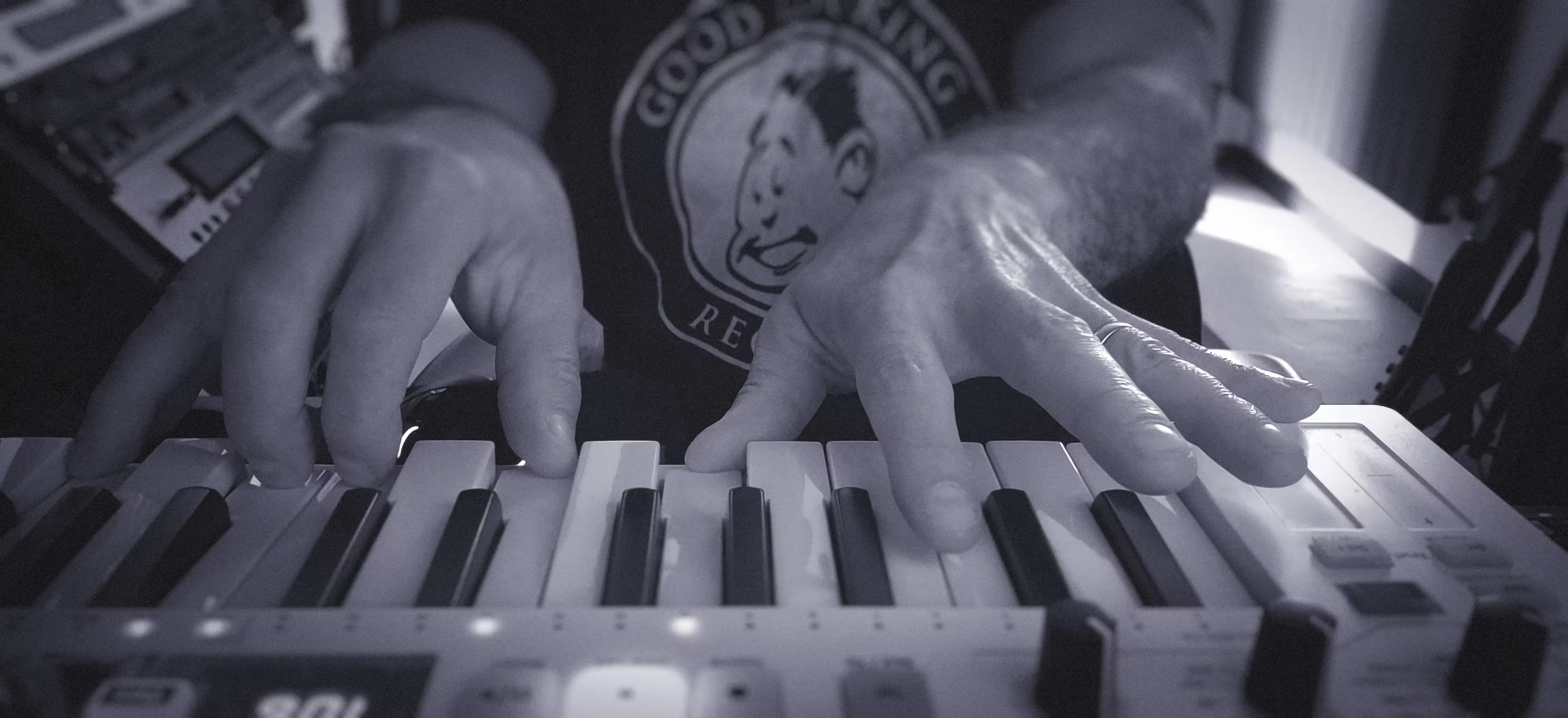 Close-up of a person playing a keyboard synthesizer with both hands, wearing a "Good Looking Records" t-shirt, in a dimly lit room.