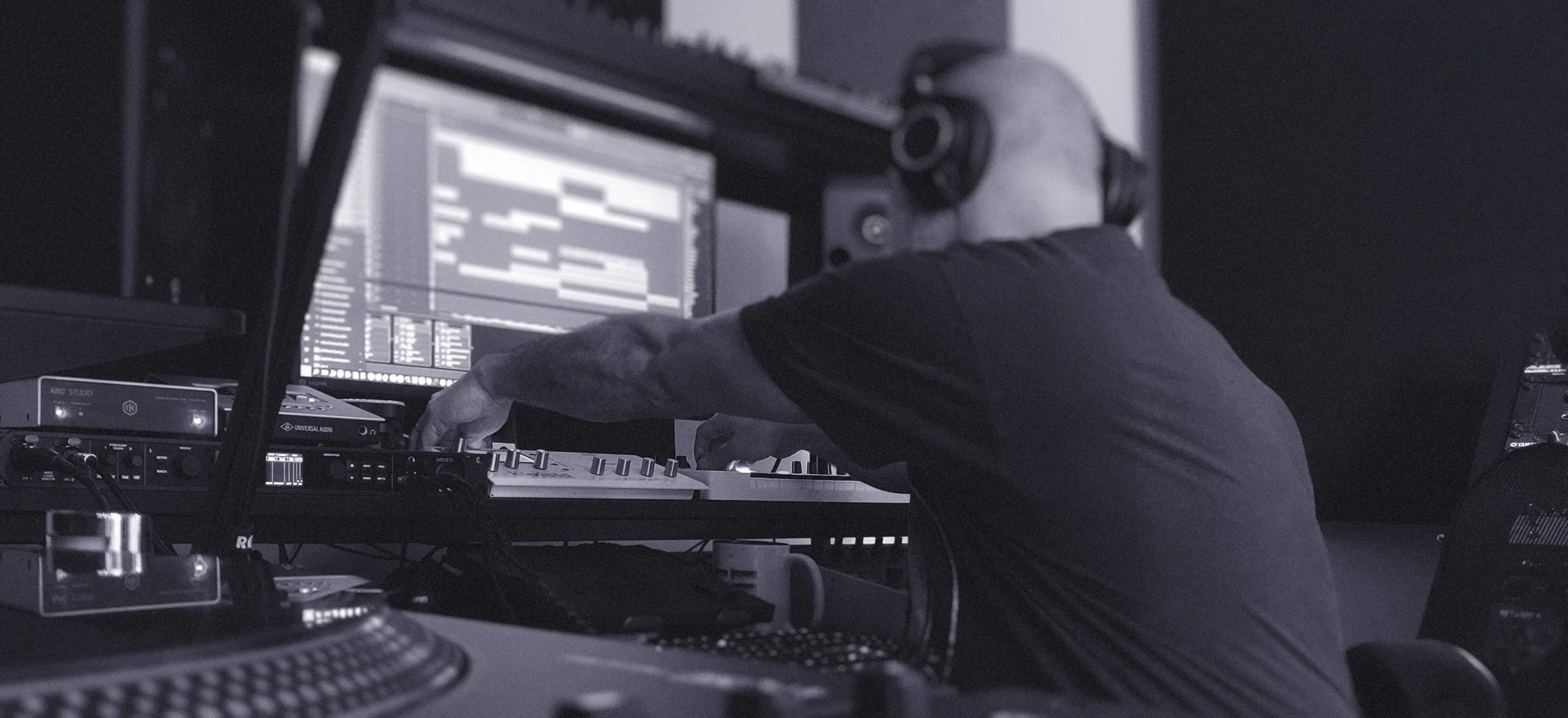 A producer wearing headphones sits at a music production desk, reaching toward a keyboard controller beneath a large computer monitor displaying audio editing software. He is in the process of converting AI to human music recreation. The studio includes speakers and equipment, with a turntable visible in the foreground.