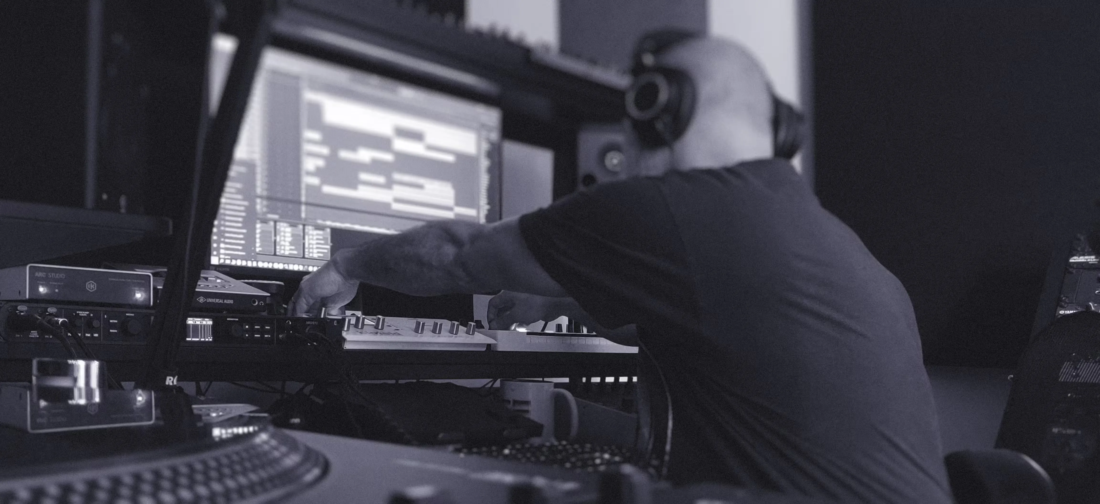 A producer wearing headphones sits at a music production desk, reaching toward a keyboard controller beneath a large computer monitor displaying audio editing software. He is in the process of converting AI to human music recreation. The studio includes speakers and equipment, with a turntable visible in the foreground.
