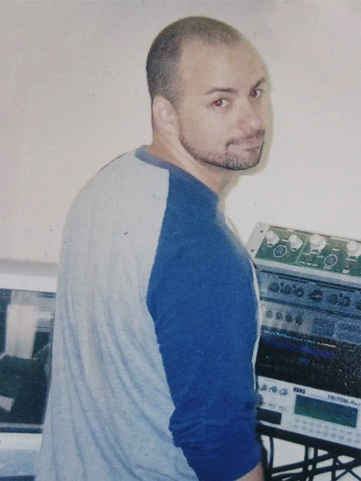 A man with a shaved head and trimmed beard, wearing a gray and blue shirt, stands in front of electronic audio equipment, looking back over his shoulder.