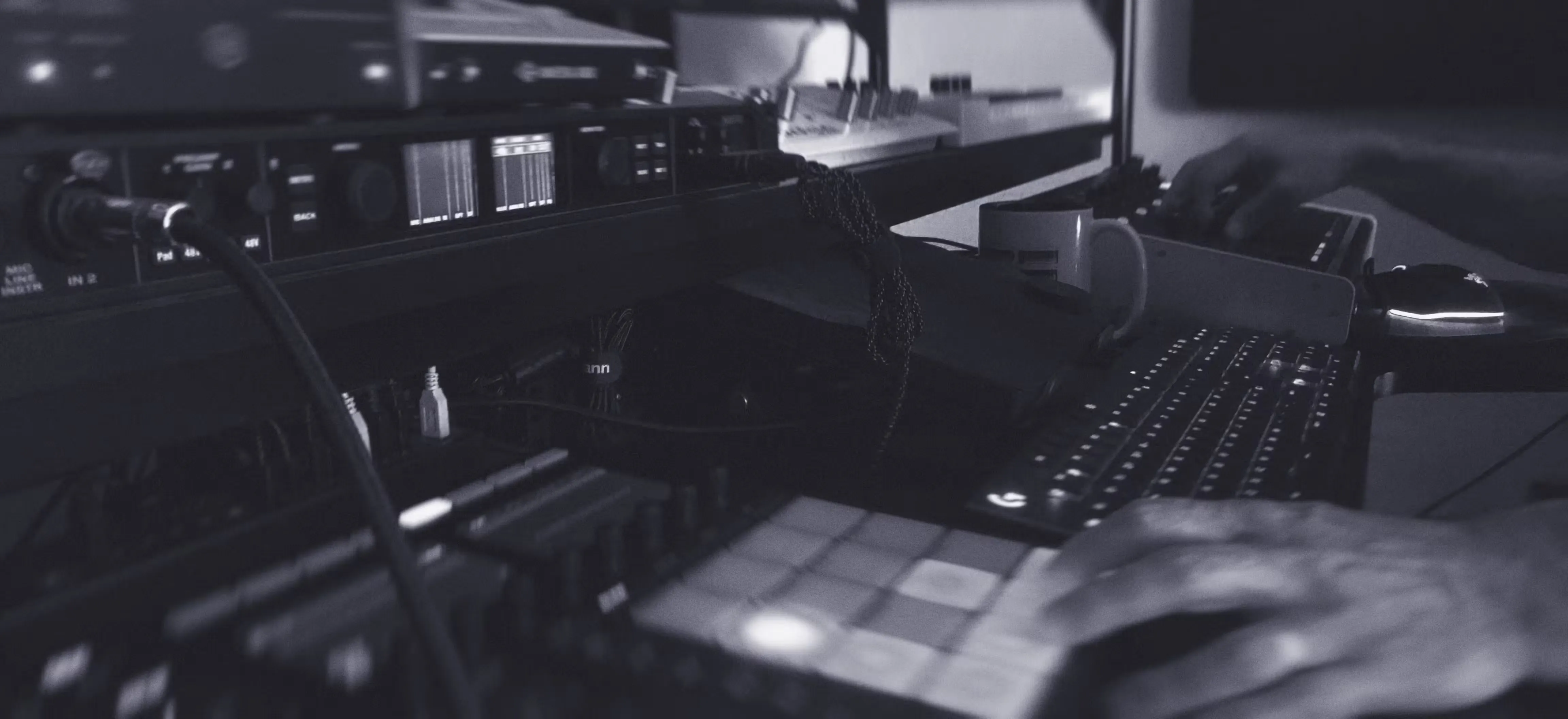 A person’s hands operate music production equipment, including a MIDI controller with pads, an audio interface, and a computer keyboard in a studio setting.