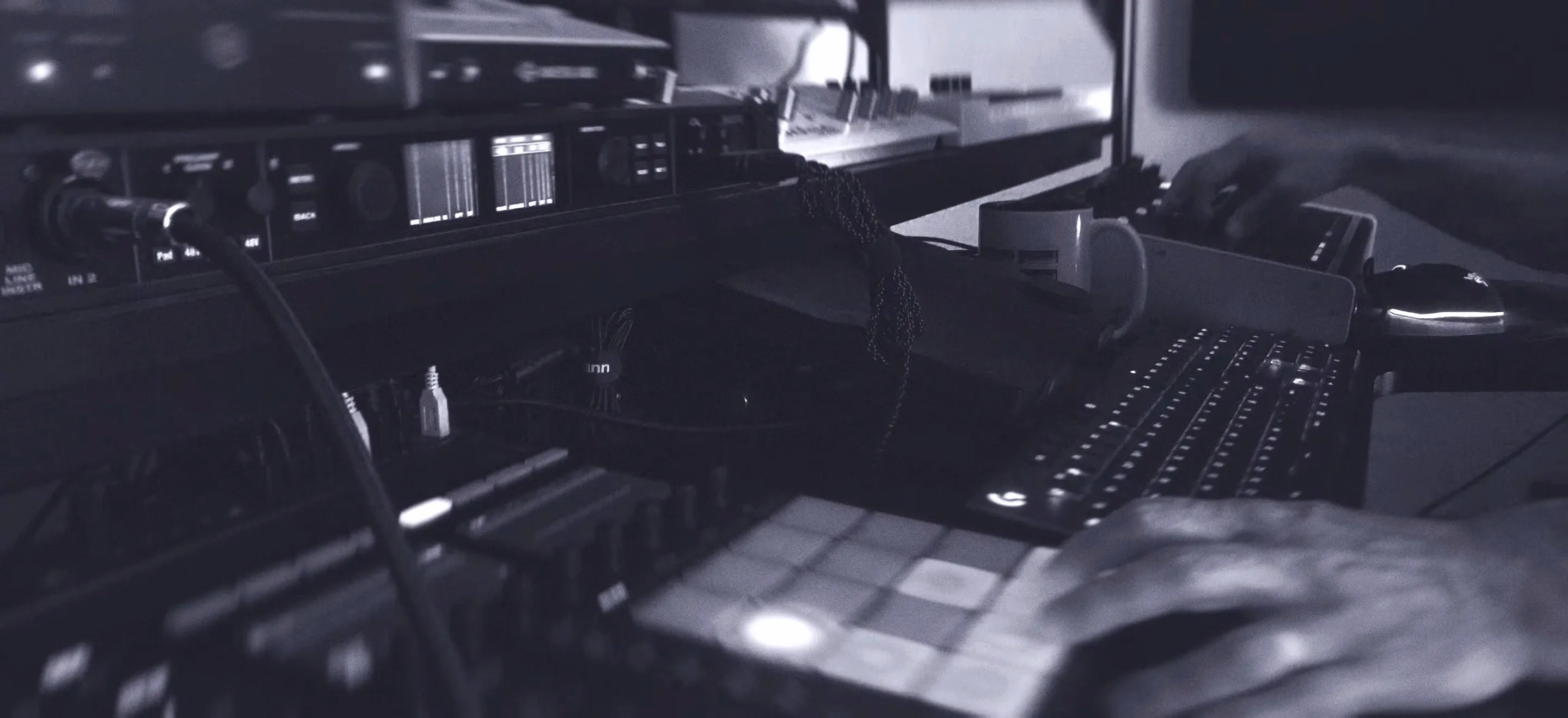 A person’s hands operate music production equipment, including a MIDI controller with pads, an audio interface, and a computer keyboard in a studio setting.