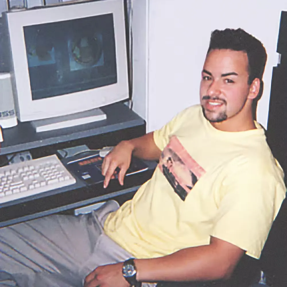 A man in a yellow T-shirt sits at a desk with an old computer and keyboard, smiling at the camera.