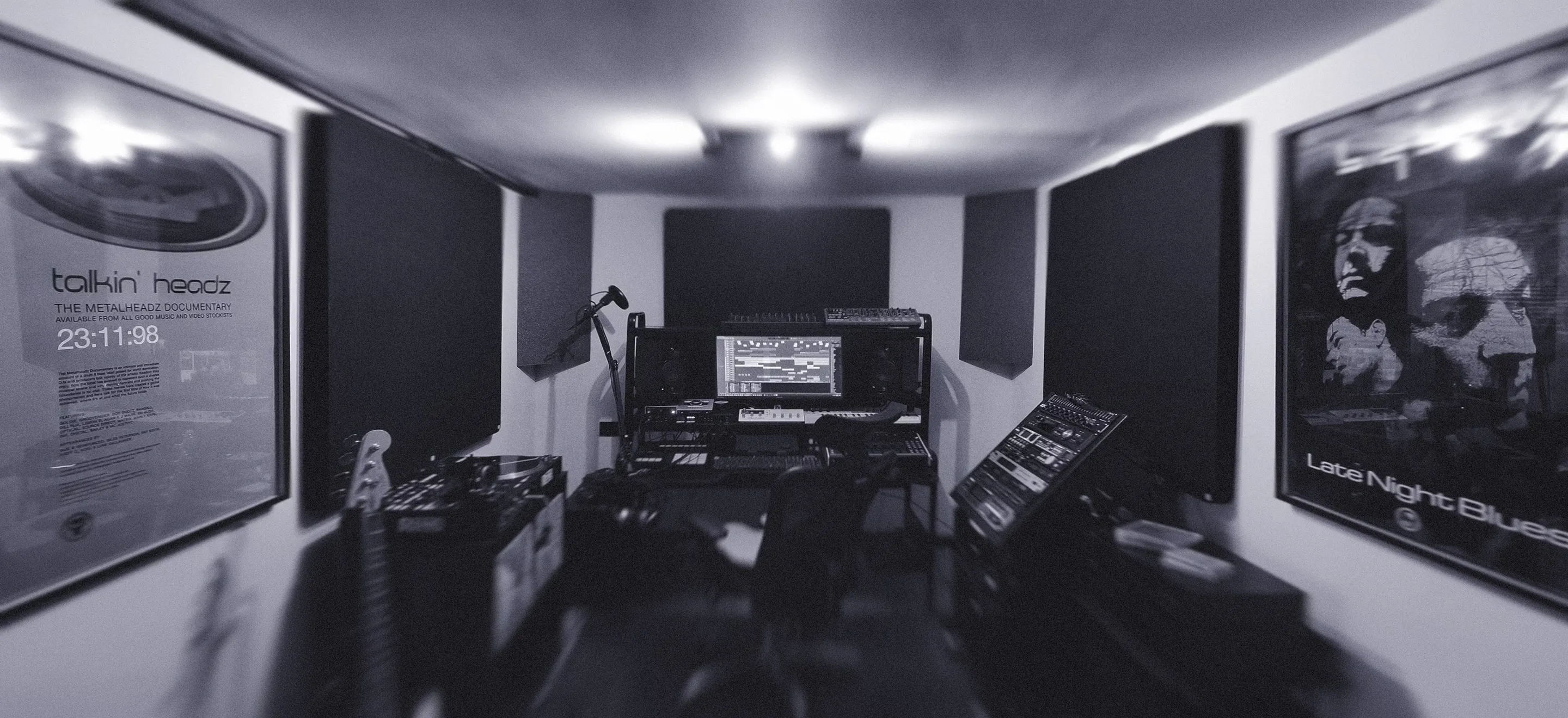 Monochrome photograph of a dimly lit music studio with sound panels, a computer on a desk, and various musical equipment. Posters adorn the walls, creating a focused yet creative vibe.