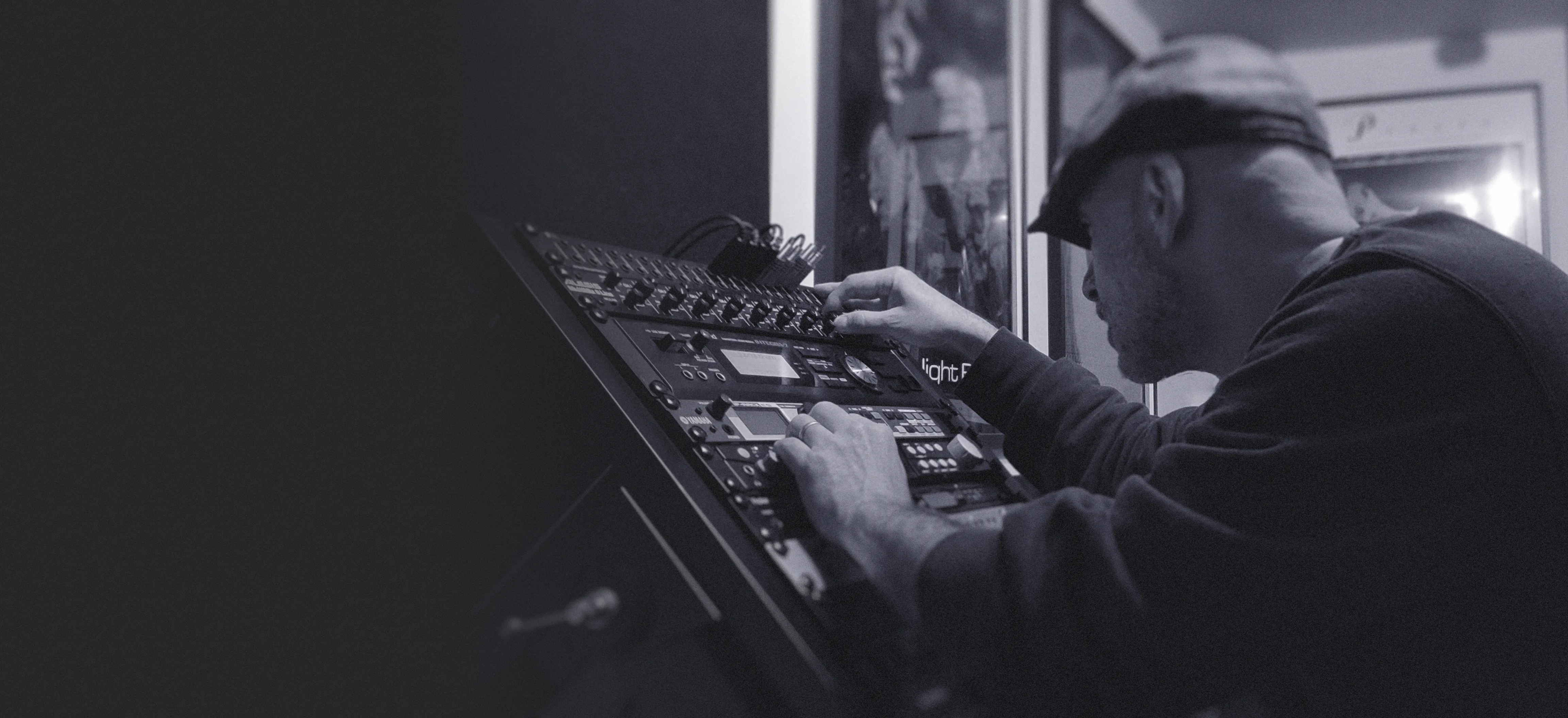 Aymat leaning into a rack of synths in his studio, adjusting knobs with both hands.