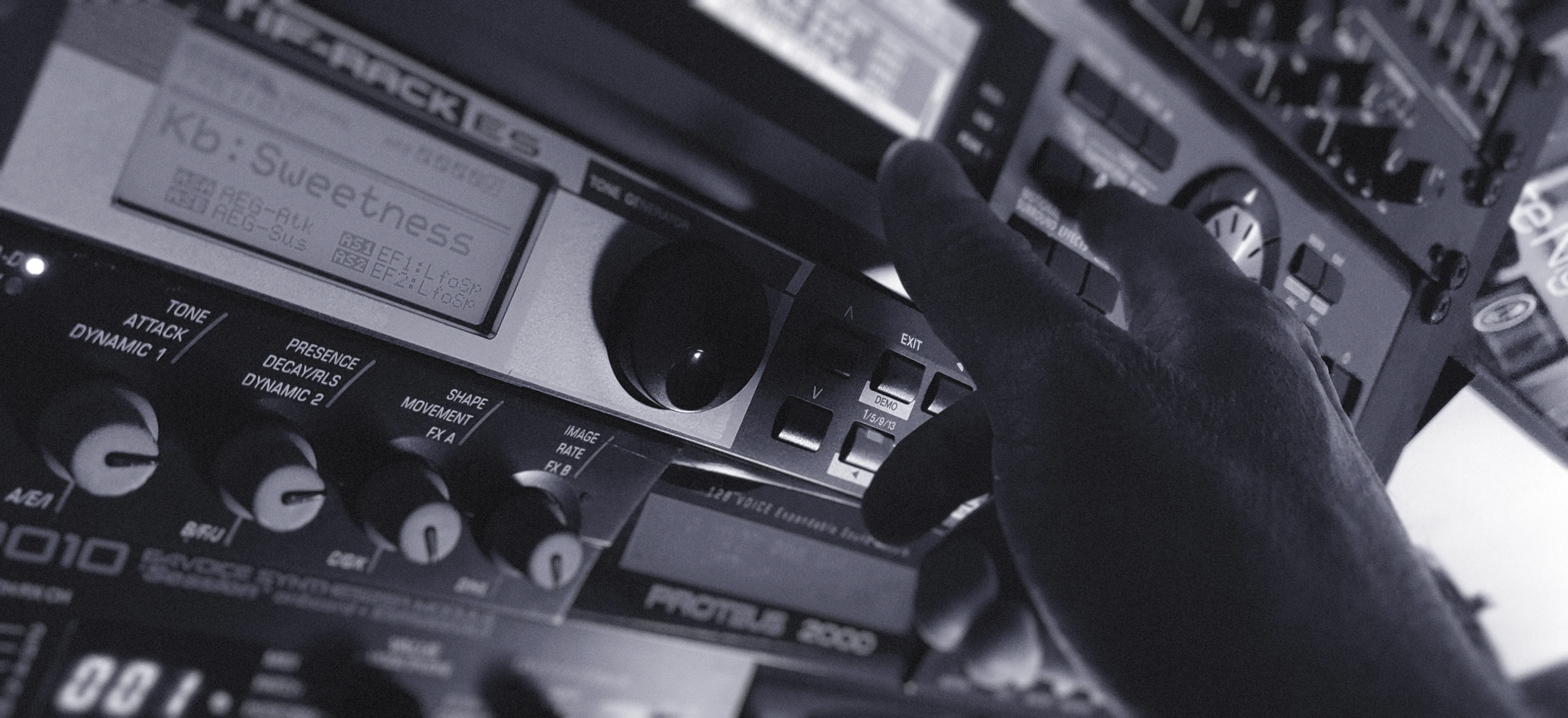 Close-up of a hand on a rack of synth modules, including a Yamaha Motif-Rack ES and an E-MU Proteus 2000.