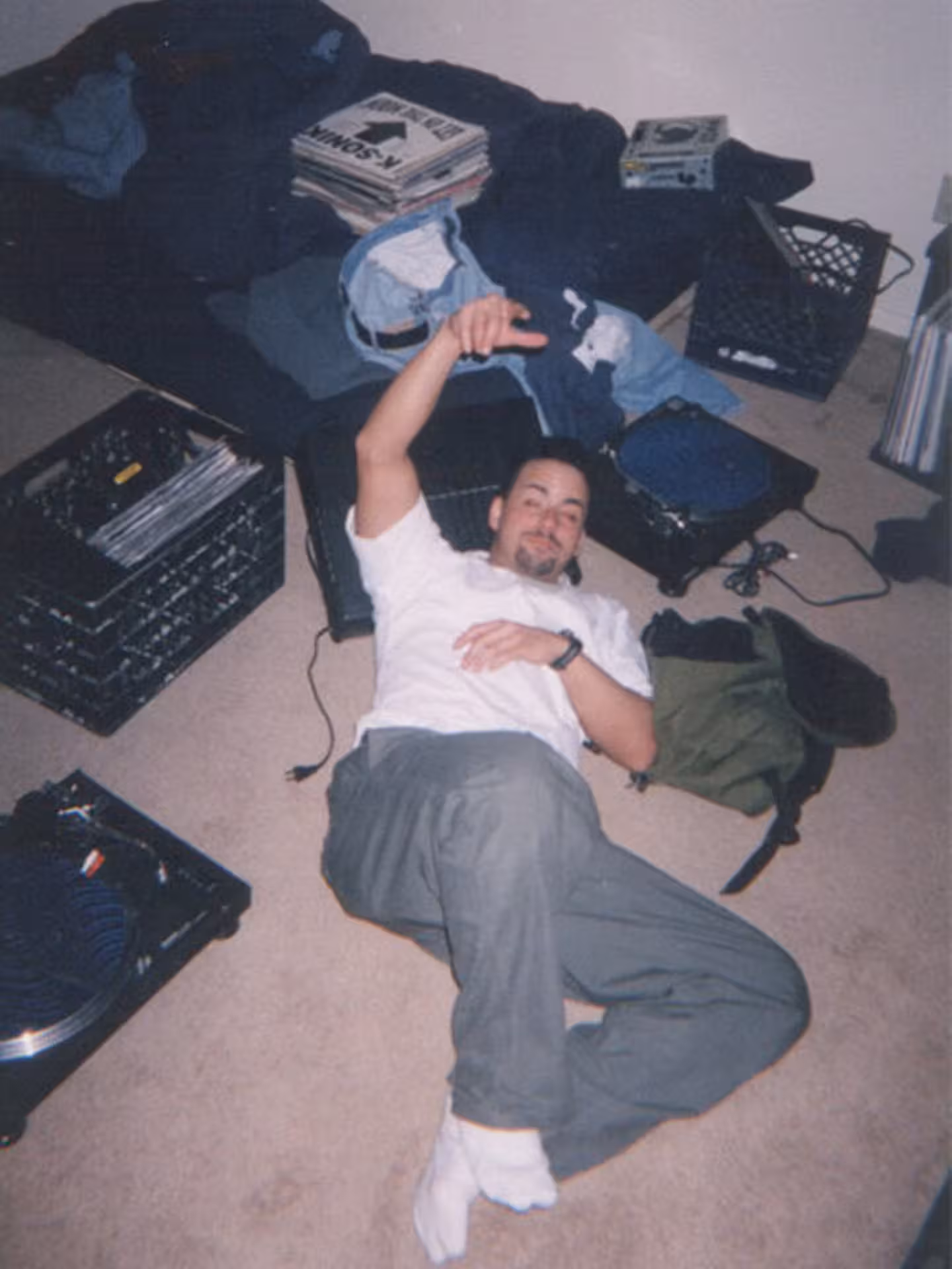 aymat-djing-1999 A man lies on the carpeted floor, surrounded by vinyl records, a turntable, milk crates, and an unmade bed in a messy bedroom.
