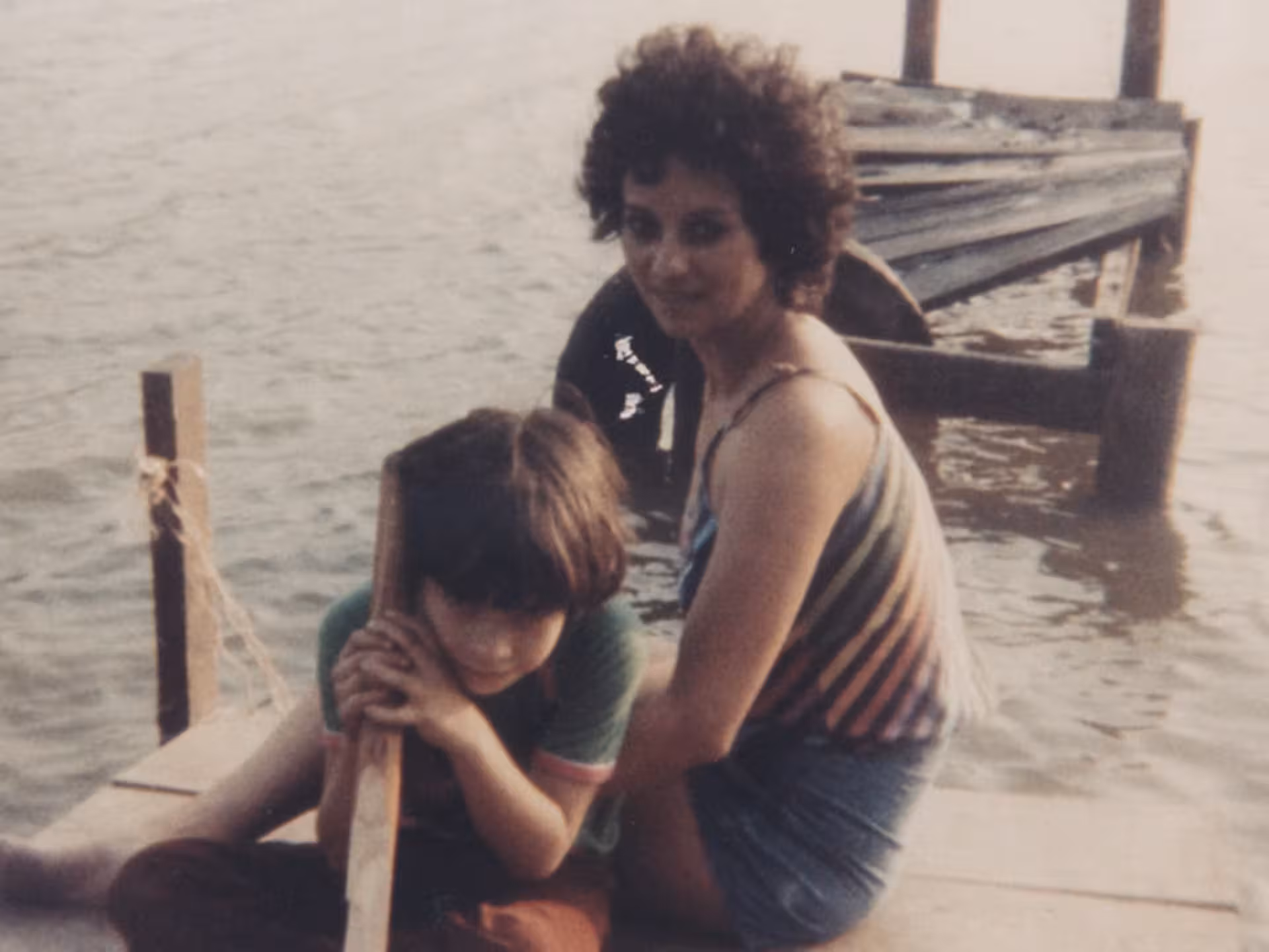 aymat-early-days A woman and a child sit on a wooden dock by the water. The woman is wearing a striped top and shorts, while the child leans on a wooden post. A damaged dock is visible in the background.