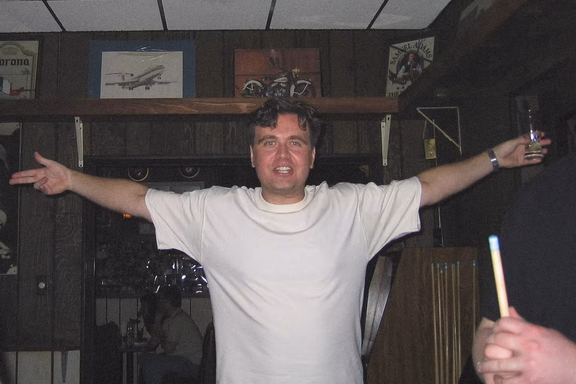 A smiling man stands in a dimly lit bar with his arms open wide. He wears a white shirt, exuding a welcoming and cheerful vibe.