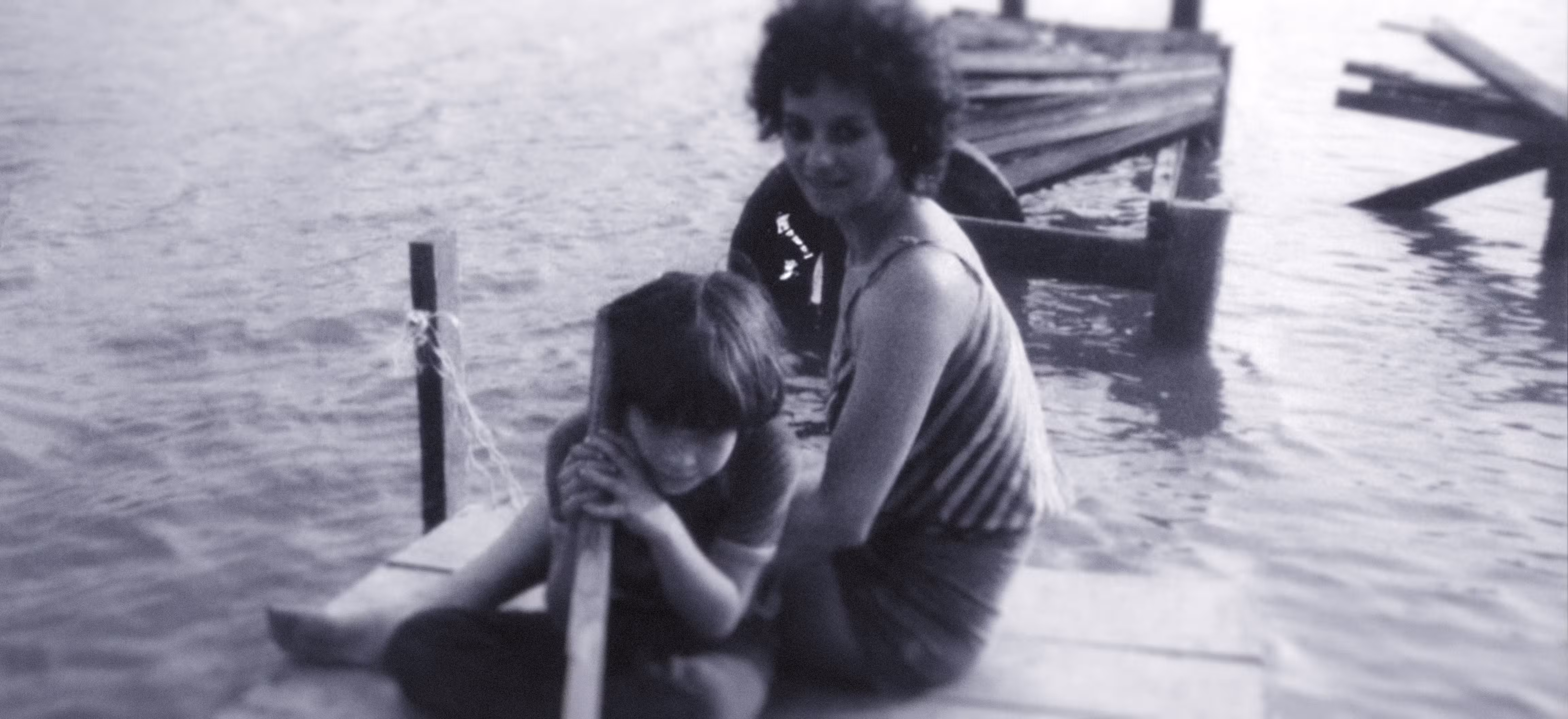 Monochrome photograph of a woman and a child sit on a wooden dock by the water; the child is holding a pole while the woman sits behind, both facing the camera. The image is for an article about a music producer journey.