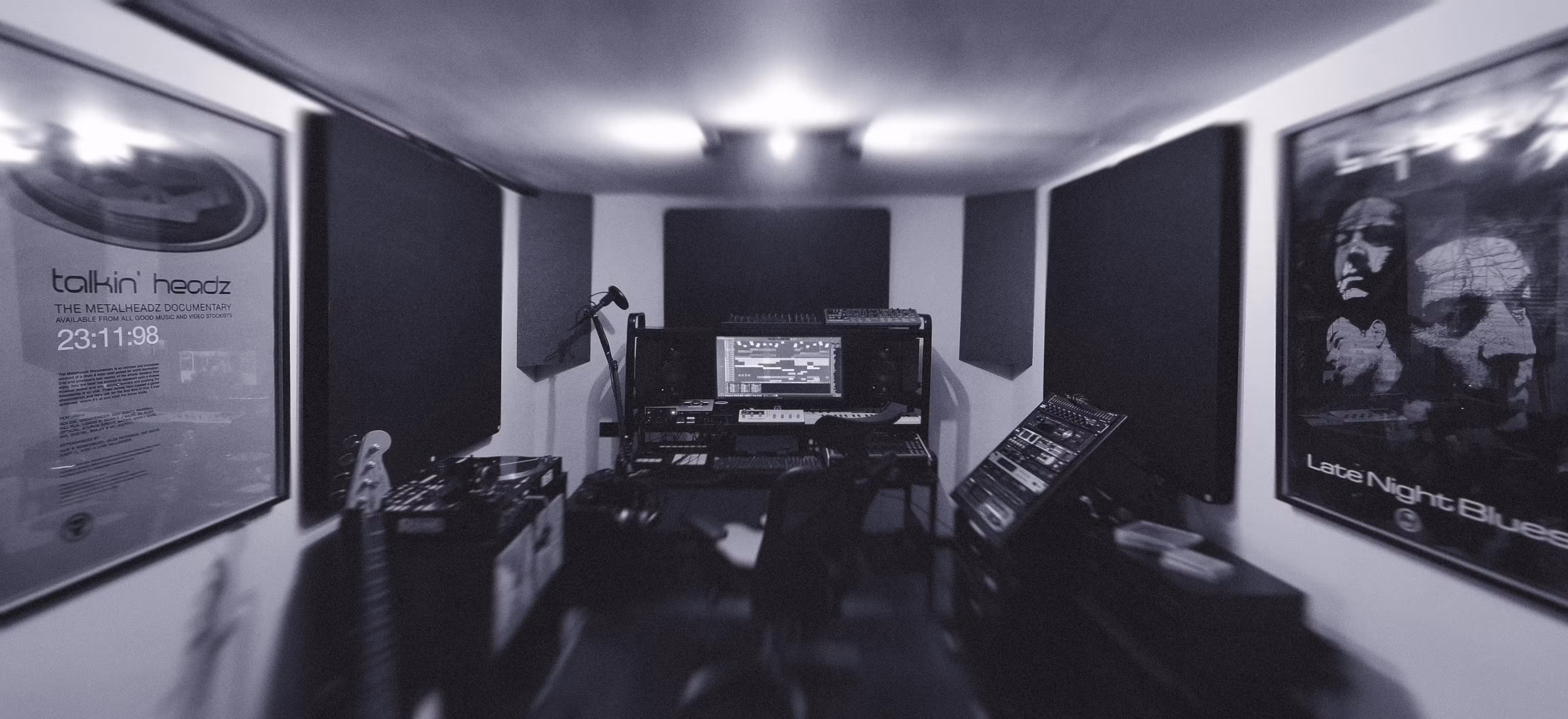 Monochrome photograph of a dimly lit music studio with sound panels, a computer on a desk, and various musical equipment. Posters adorn the walls, creating a focused yet creative vibe. The image is for an article about why most producers never finish tracks.