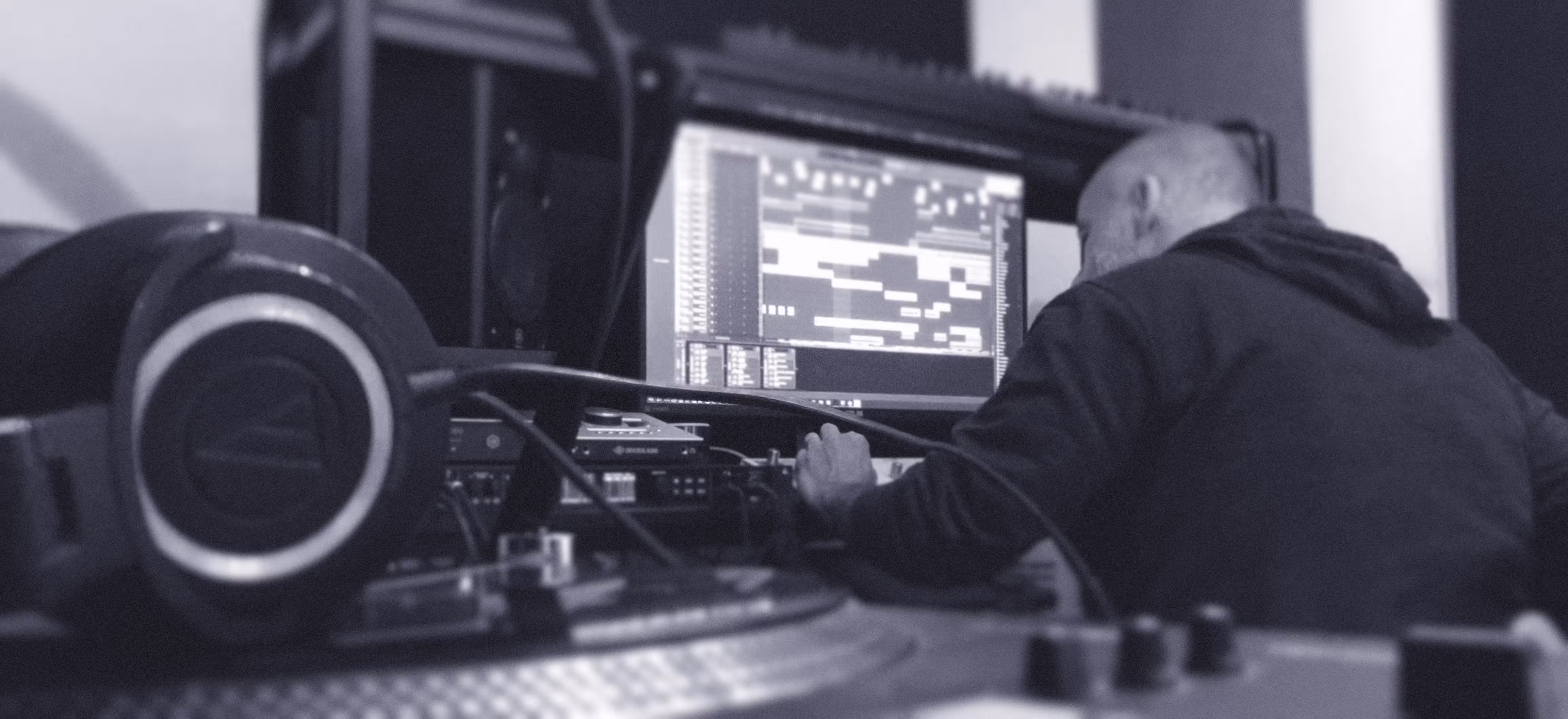 Monochrome photograph of a man working at a music production studio desk with headphones, mixer, and DAW software on screen. The image is for an article about the psychology of being a music producer.