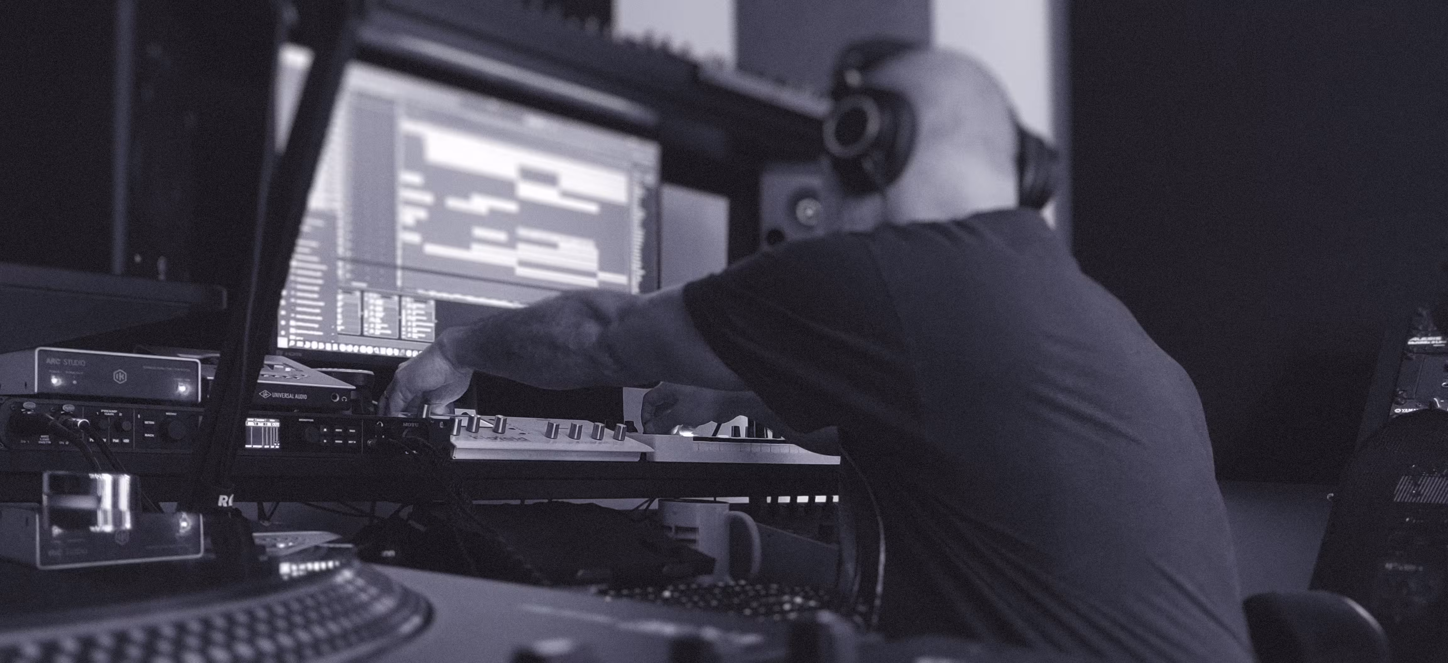 aymat-studio-004 A producer wearing headphones sits at a music production desk, reaching toward a keyboard controller beneath a large computer monitor displaying audio editing software. He is in the process of converting AI to human music recreation. The studio includes speakers and equipment, with a turntable visible in the foreground.