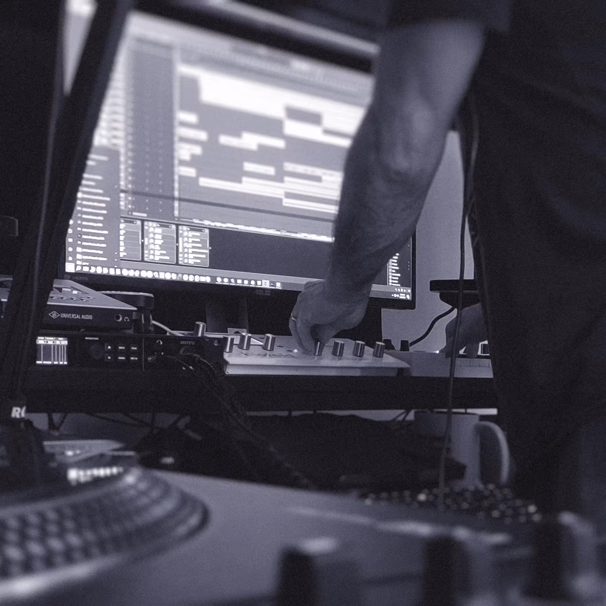 A person using music production equipment in a studio setting, with computer screens displaying software and audio tracks in the background.