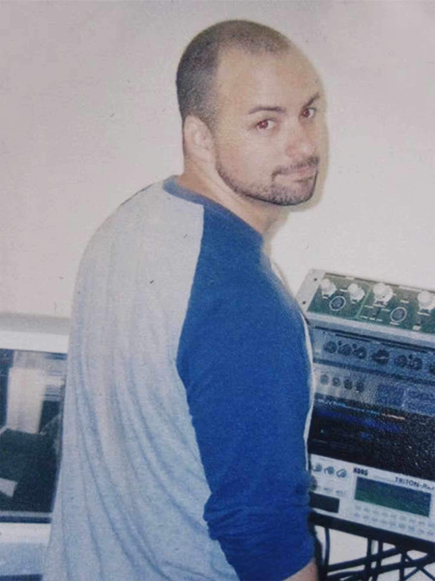 aymat-studio-2004 A man with a shaved head and trimmed beard, wearing a gray and blue shirt, stands in front of electronic audio equipment, looking back over his shoulder.