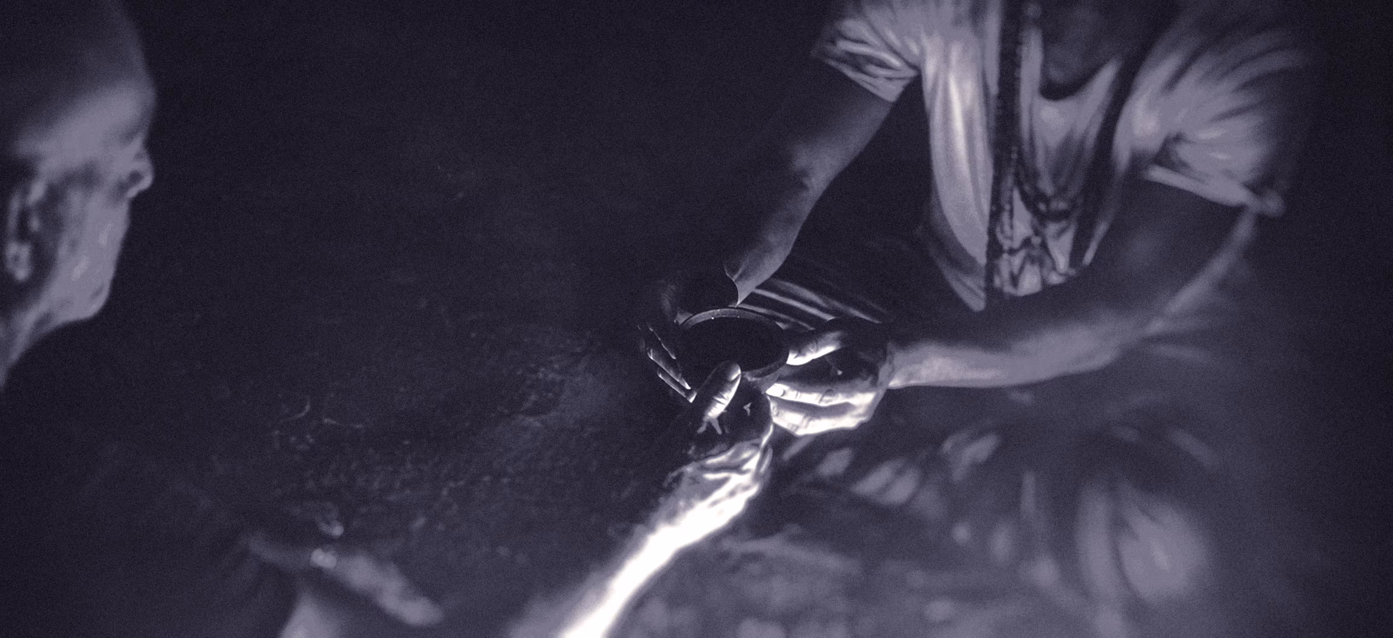 Two people are sitting in low light at an ayahuasca ritual, with one person handing a small round bowl, to the other. The image is for an article about self-doubt as a music producer.