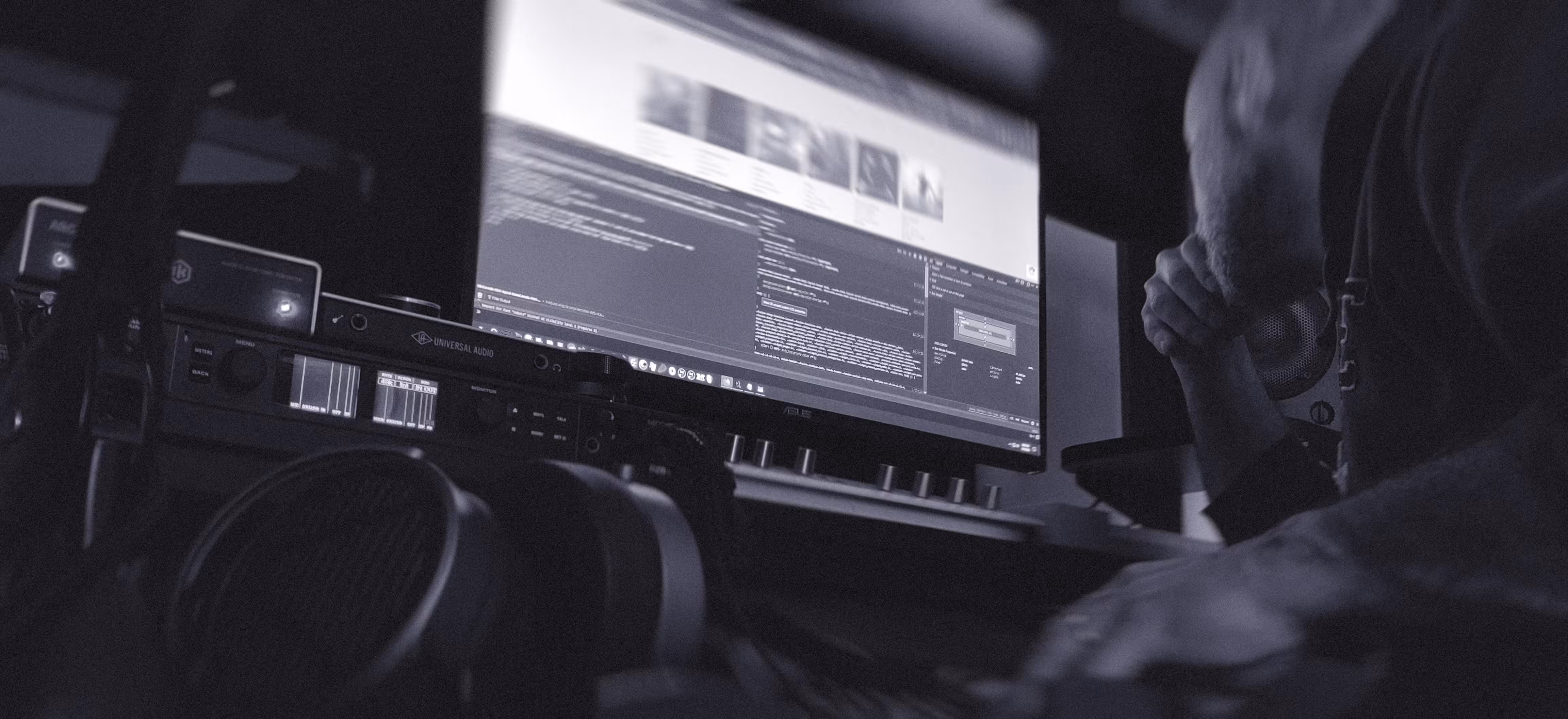 A person leans over a desk, surrounded by audio equipment, looking at a large computer monitor displaying code and images in a dimly lit workspace, capturing a moment of creative development for music artists