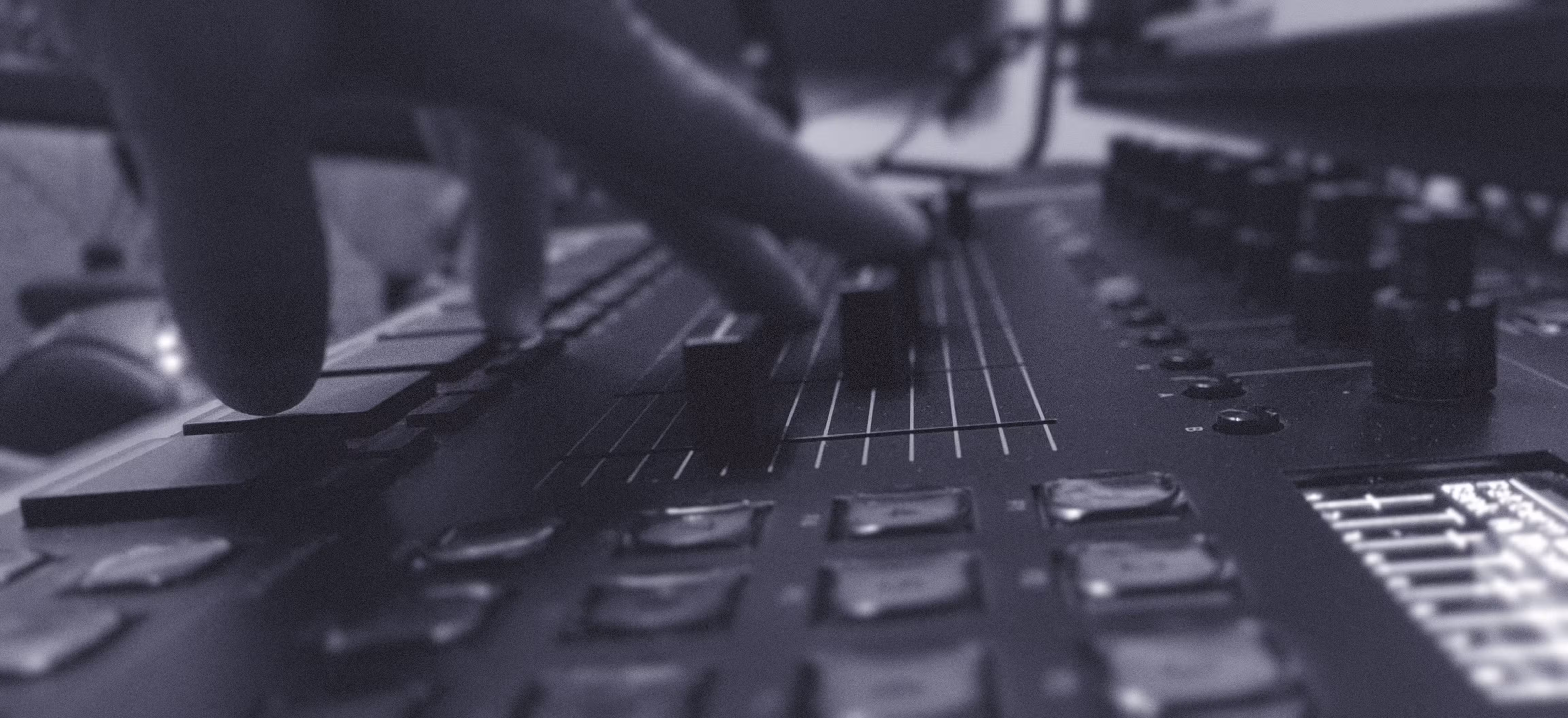 A producer coaching with hand adjusting sliders on a sound mixing console, control knobs and buttons are visible in the foreground.