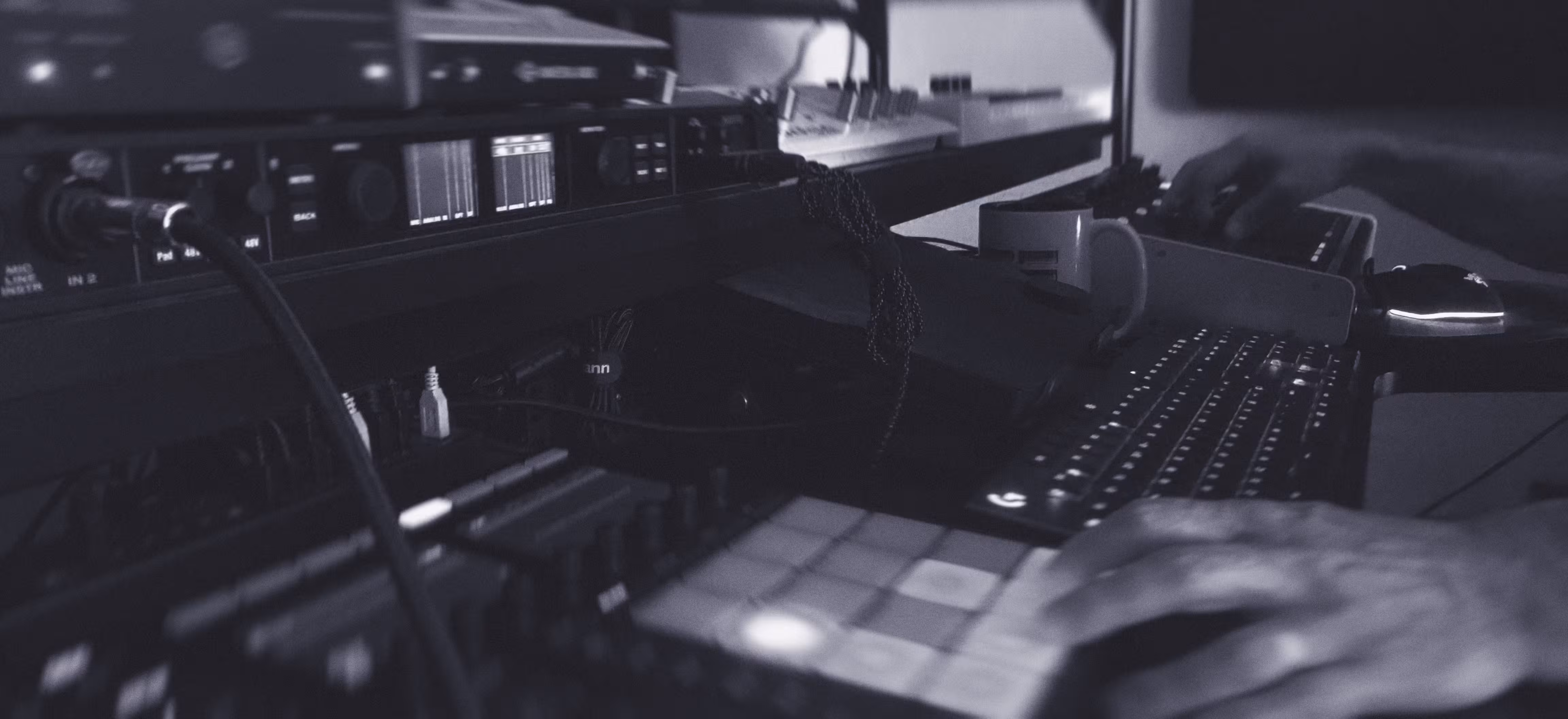 A person’s hands operate music production equipment, including a MIDI controller with pads, an audio interface, and a computer keyboard in a studio setting.