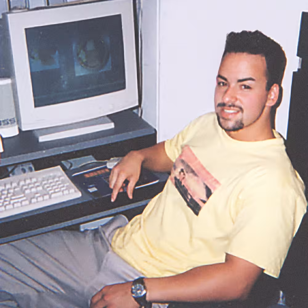 A man in a yellow T-shirt sits at a desk with an old computer and keyboard, smiling at the camera.